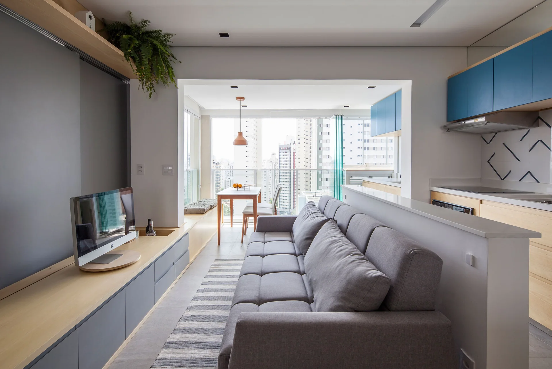 A grey sofa sits in an open-concept living area next to a low white partition, opposite a light wood media console and blue kitchen cabinets.