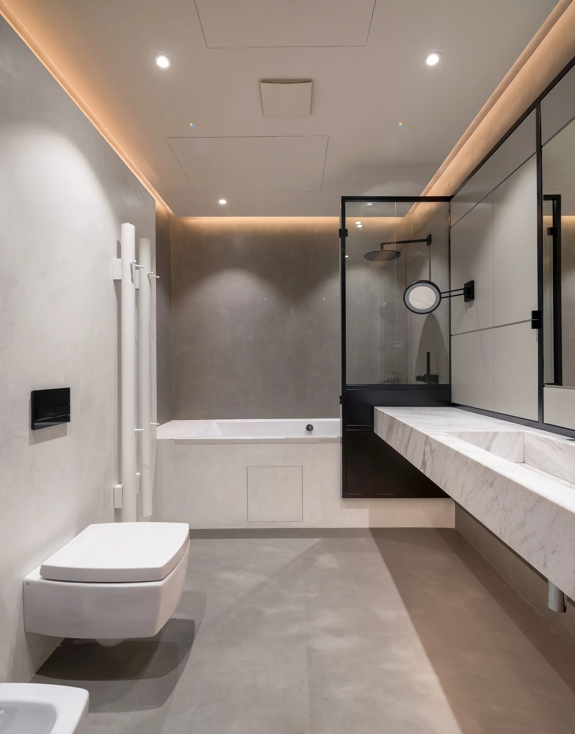 A contemporary grey bathroom featuring a floating white marble vanity, a wall-mounted toilet, a built-in bathtub, and soft ceiling cove lighting.