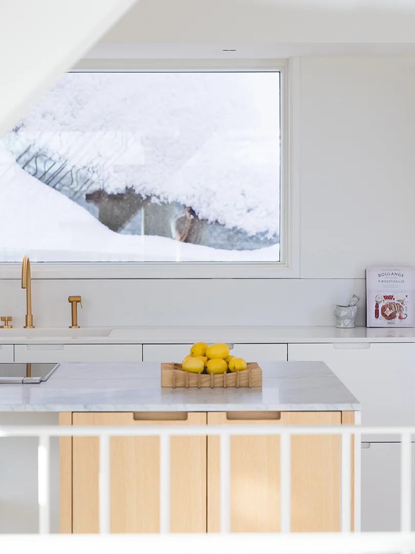 A minimalist white kitchen features a light wood island with a marble countertop, a gold faucet, and a window showing a snowy winter landscape.