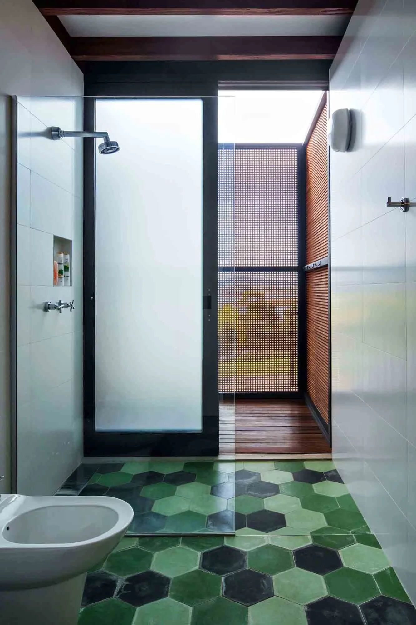 A modern bathroom features green hexagonal floor tiles, a glass shower partition, and a frosted sliding door opening to an outdoor deck with wood screening.