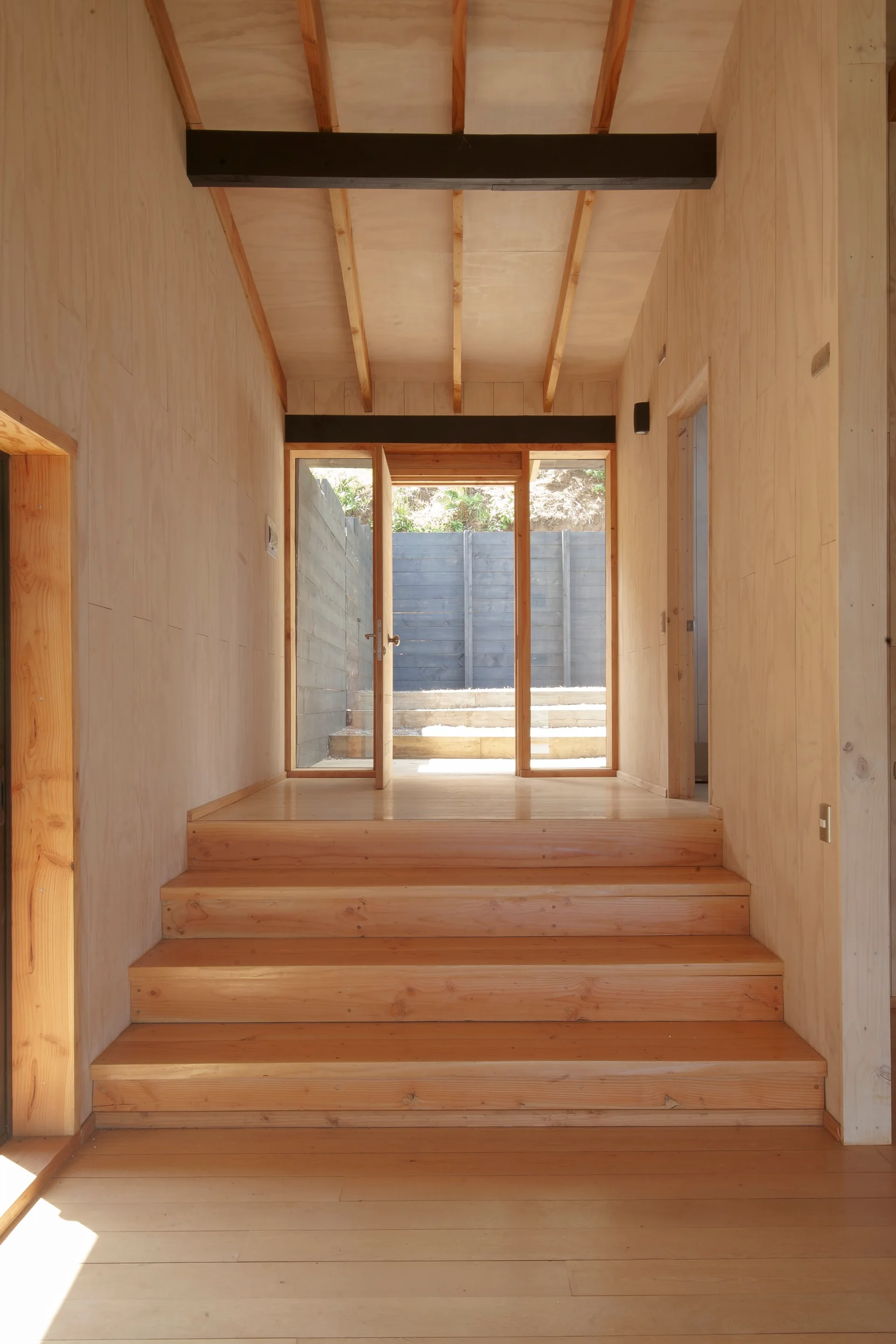 Plywood-clad walls line a wooden staircase leading up to a glass exterior door under exposed rafter ceilings with dark contrast beams.