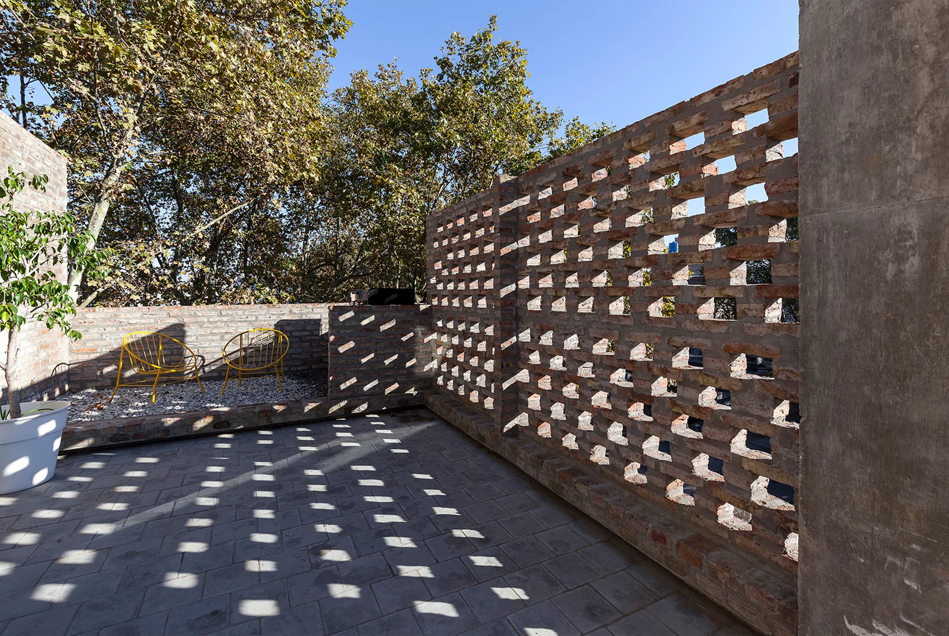A modern paved stone terrace features a perforated brick screen wall casting geometric shadow patterns, with yellow metal chairs in the background.