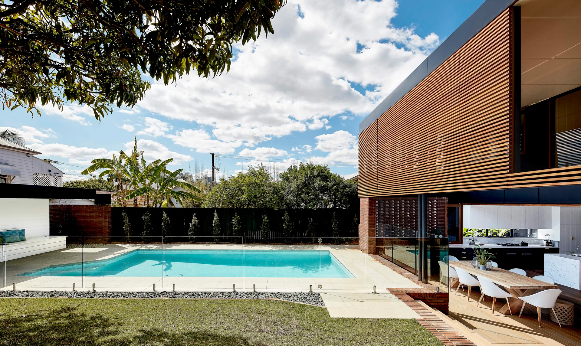 A rectangular swimming pool with a glass fence sits adjacent to a modern home featuring a timber slatted facade and an outdoor dining area.