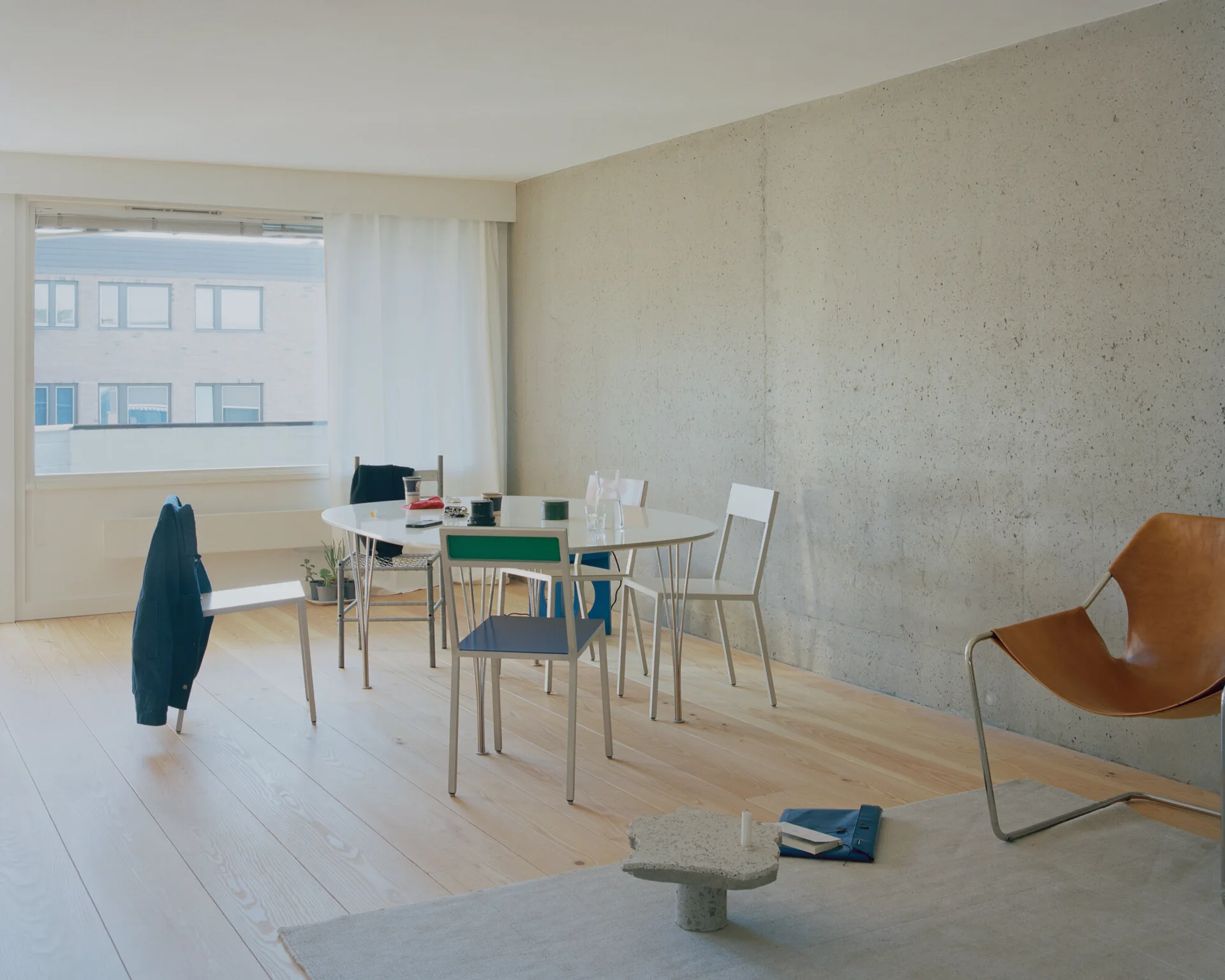 A minimalist dining room featuring an exposed concrete wall, a white oval table with metal chairs, and light wood flooring.