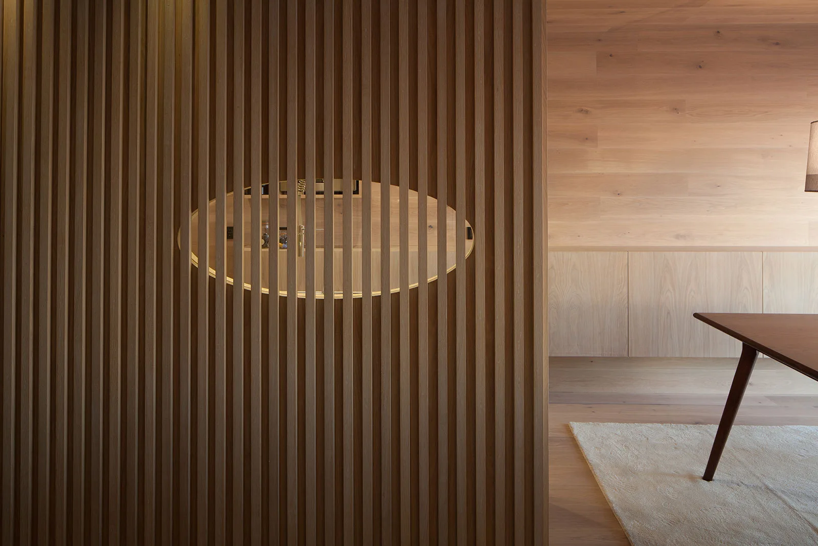 A vertical slatted wooden screen features an oval cutout showing a warm interior space alongside wood-paneled walls and a wooden table.