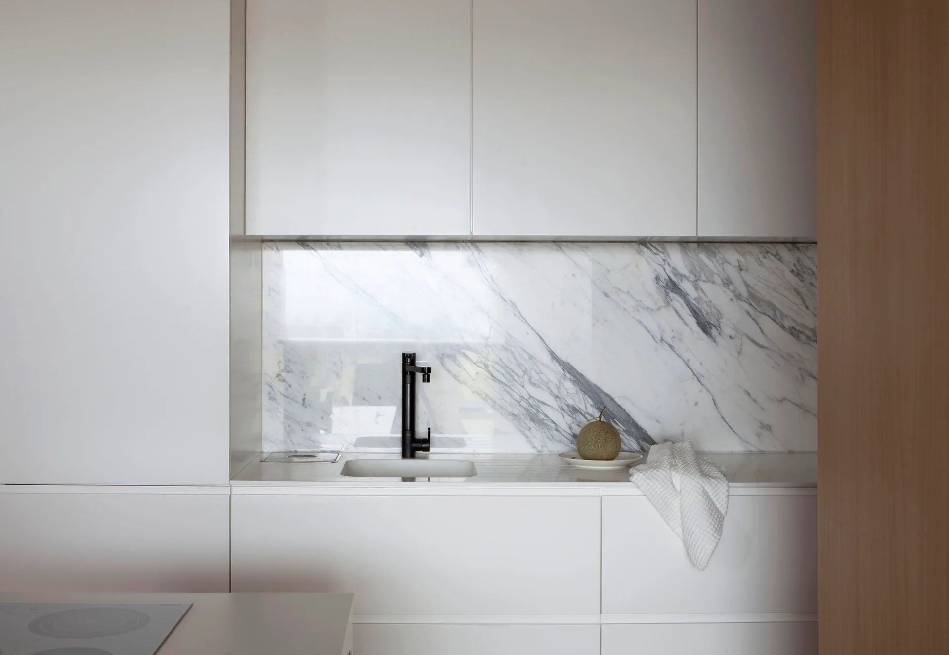 A minimalist white kitchen features handleless matte cabinetry, a heavily veined marble backsplash, a black faucet, and an integrated sink.