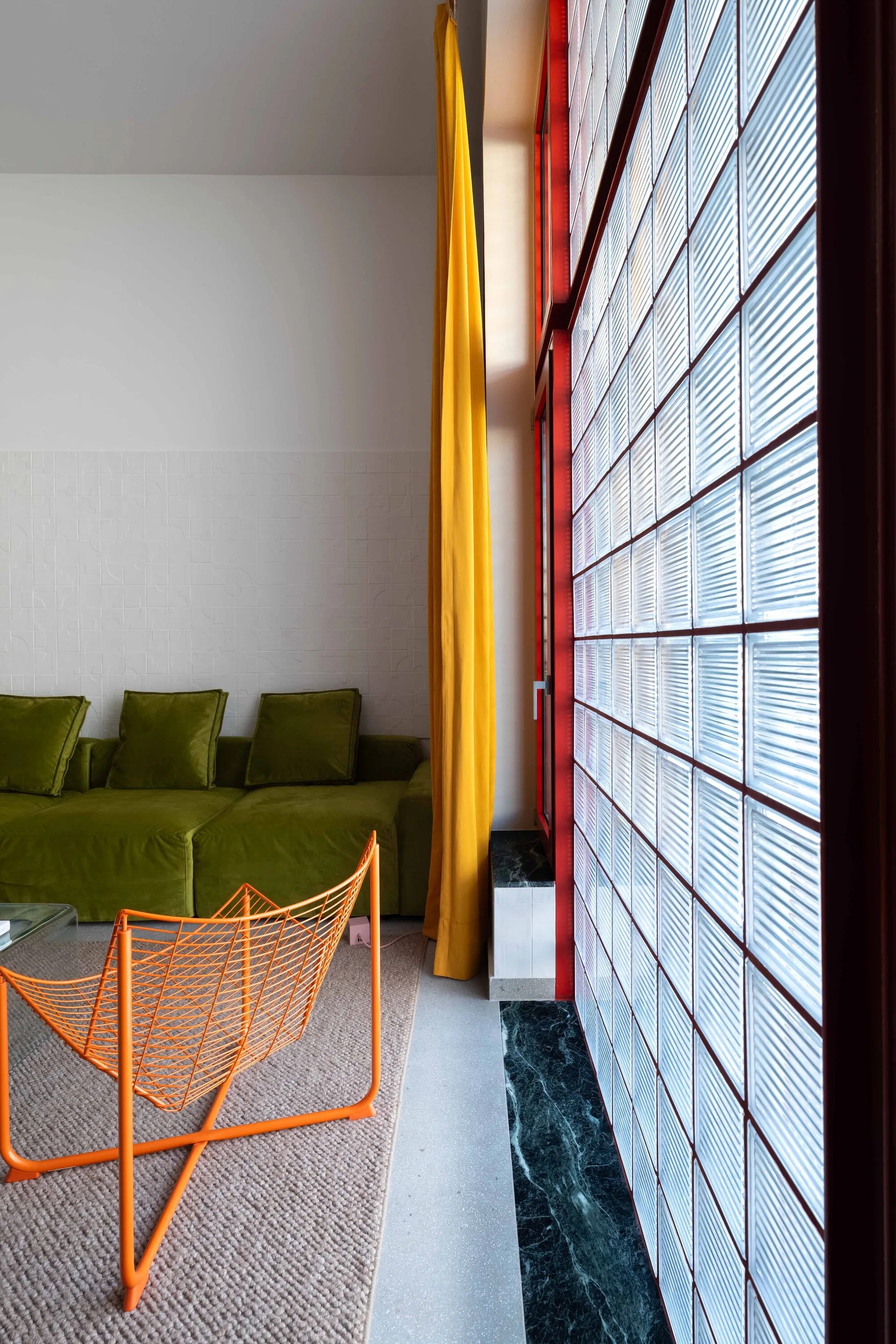 A modern living area with an olive green velvet sofa, an orange wire lounge chair, and a large red-framed glass block window.