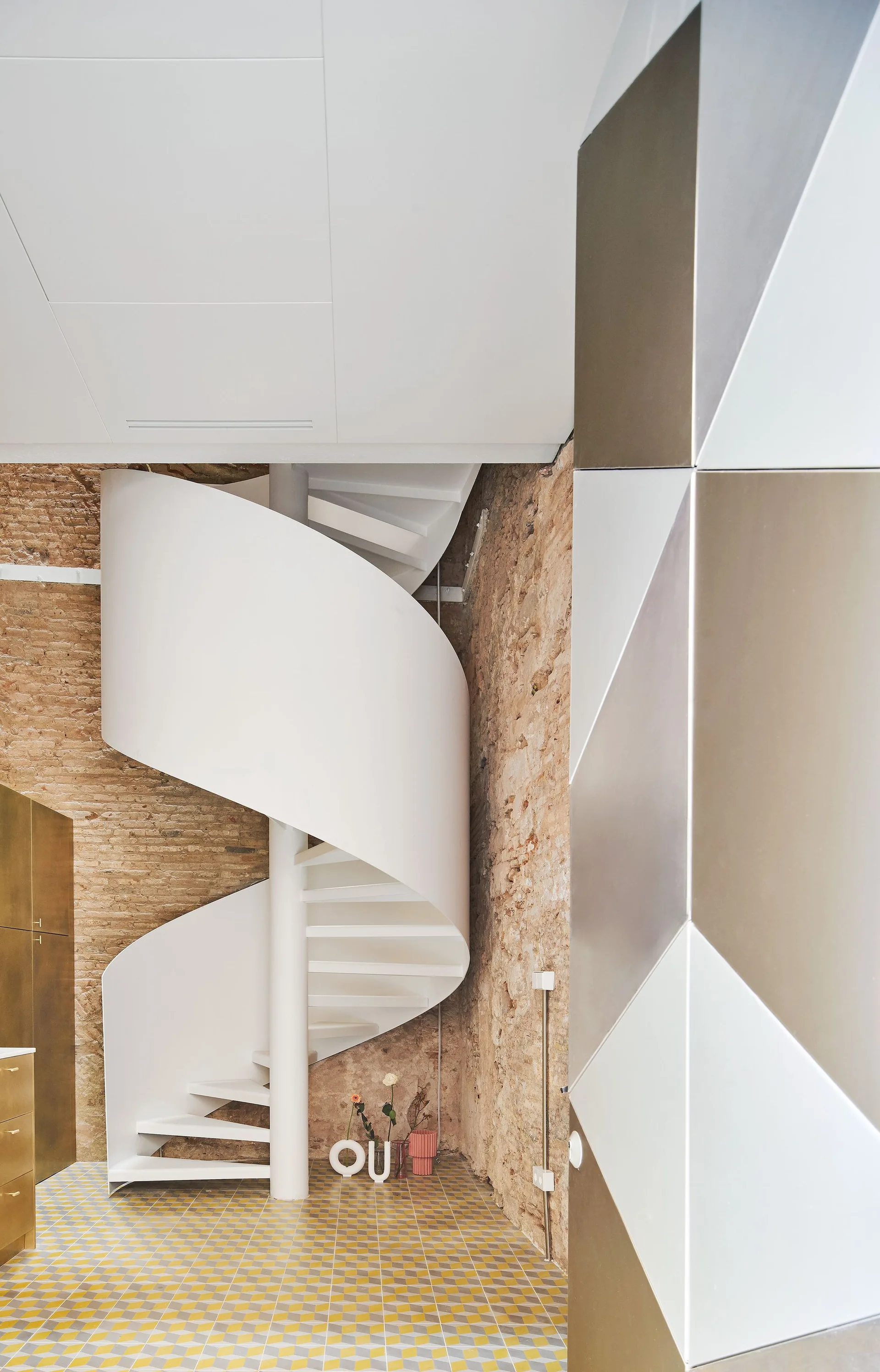 A modern white spiral staircase is set against a rustic exposed brick wall and a geometric patterned tile floor.