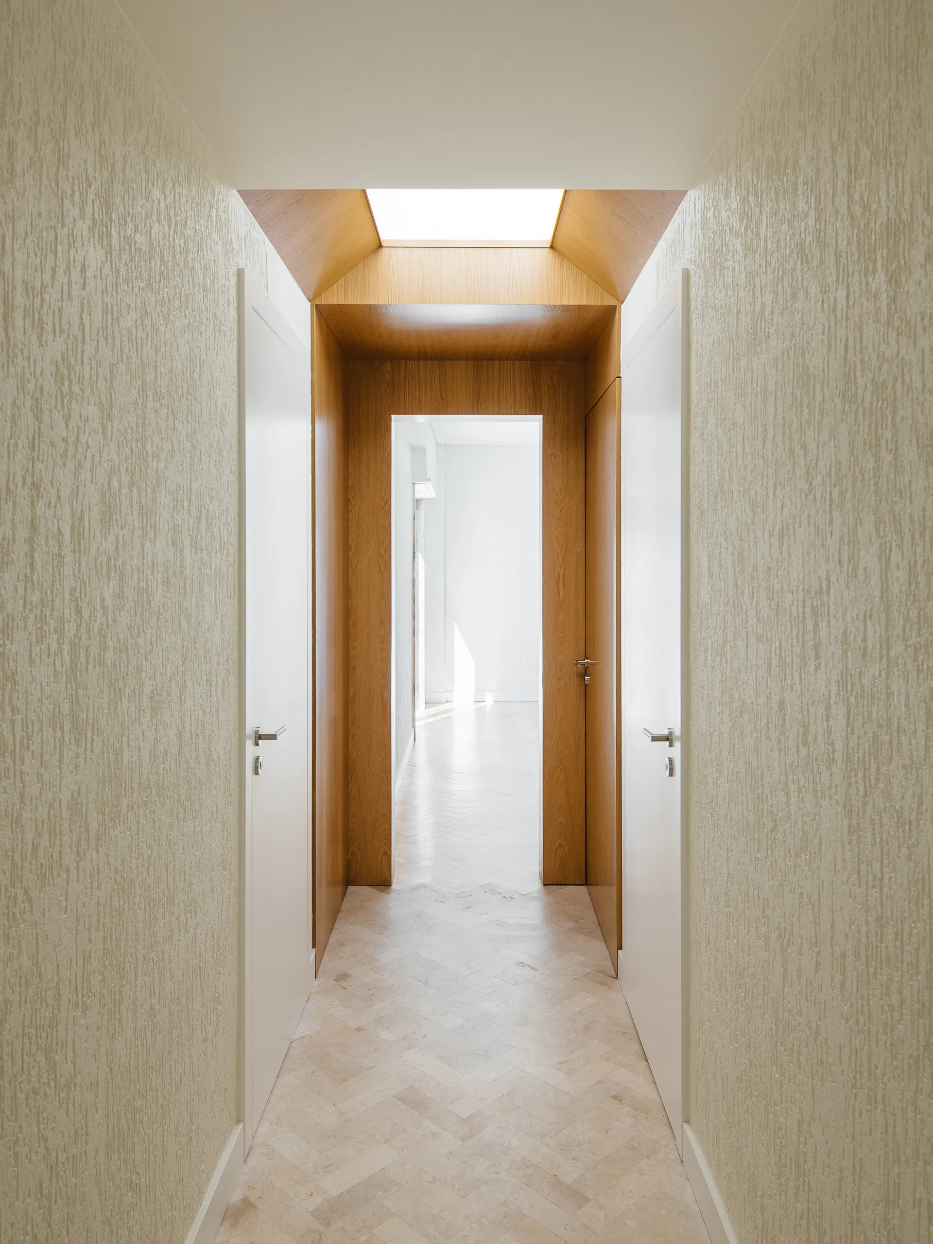 A narrow hallway features textured cream walls, herringbone stone floors, and a wood-paneled archway beneath a bright skylight.