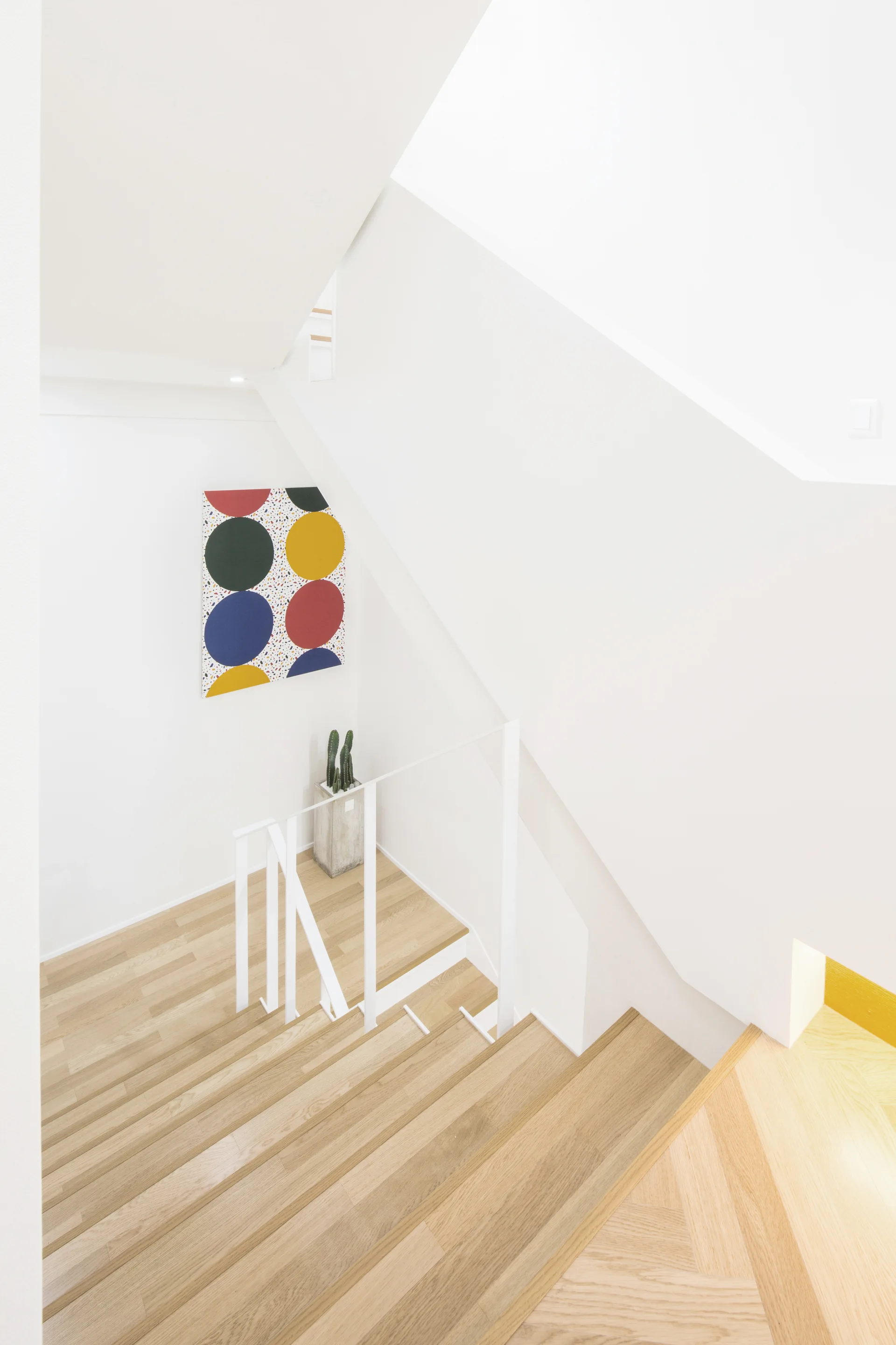 Looking down a modern staircase featuring light wood treads, white minimalist railings, a concrete planter with cacti, and colorful geometric wall art.