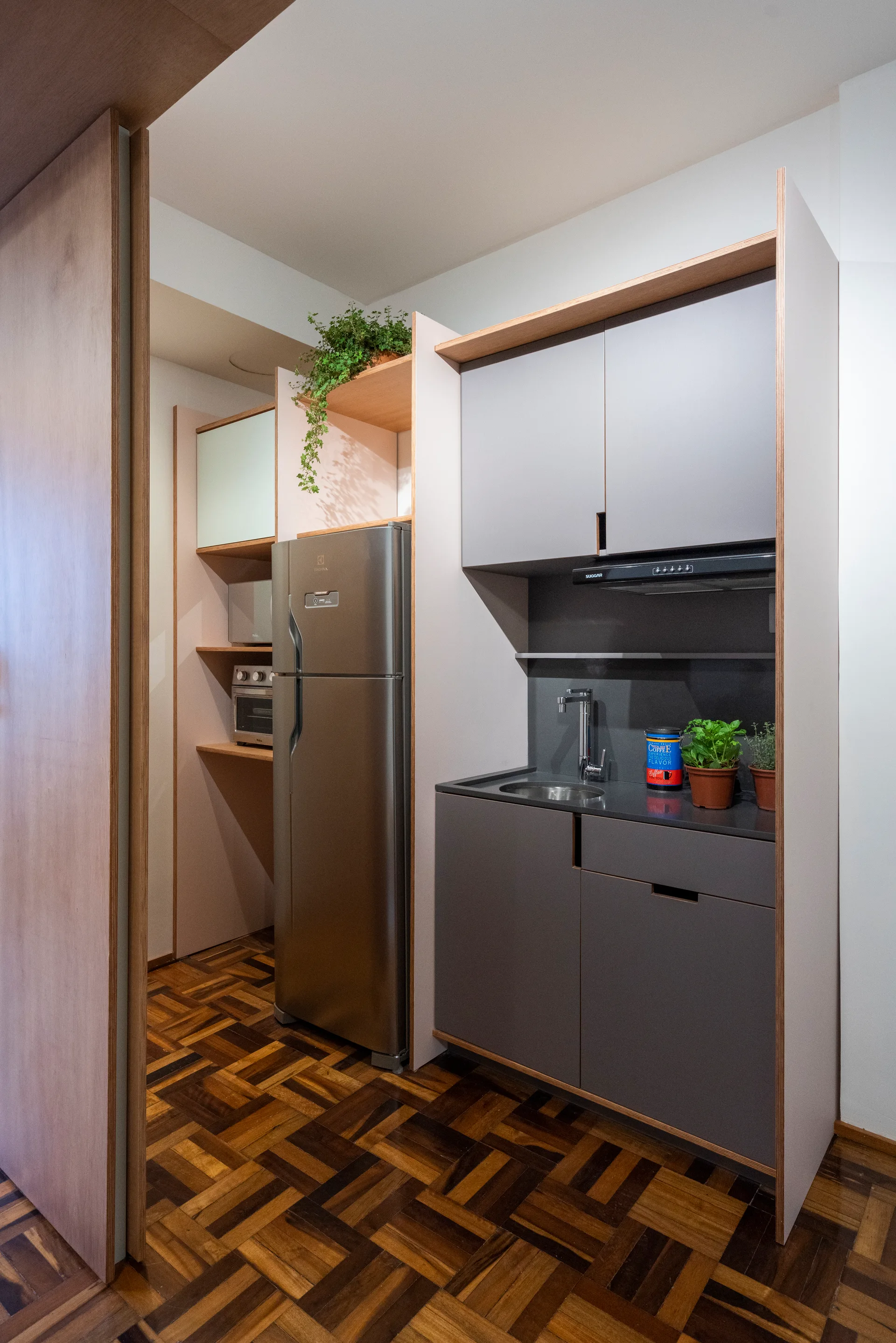 A compact kitchen features grey matte cabinets with light wood trim, a stainless steel refrigerator, and dark wood parquet block flooring.
