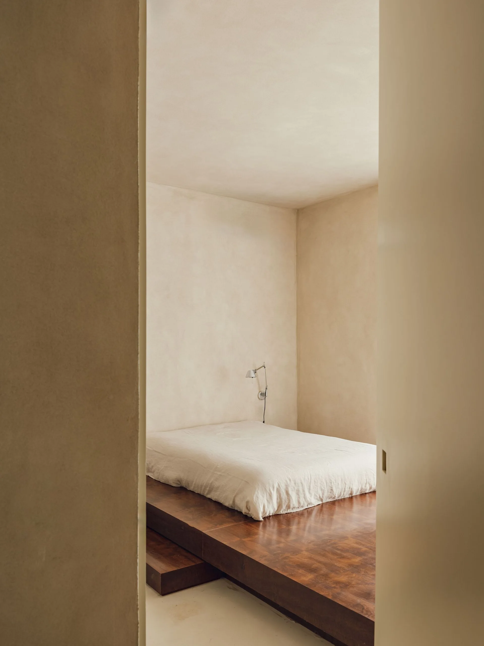 A minimalist bedroom featuring a low bed on a raised wooden platform against textured beige plaster walls with a wall-mounted light.
