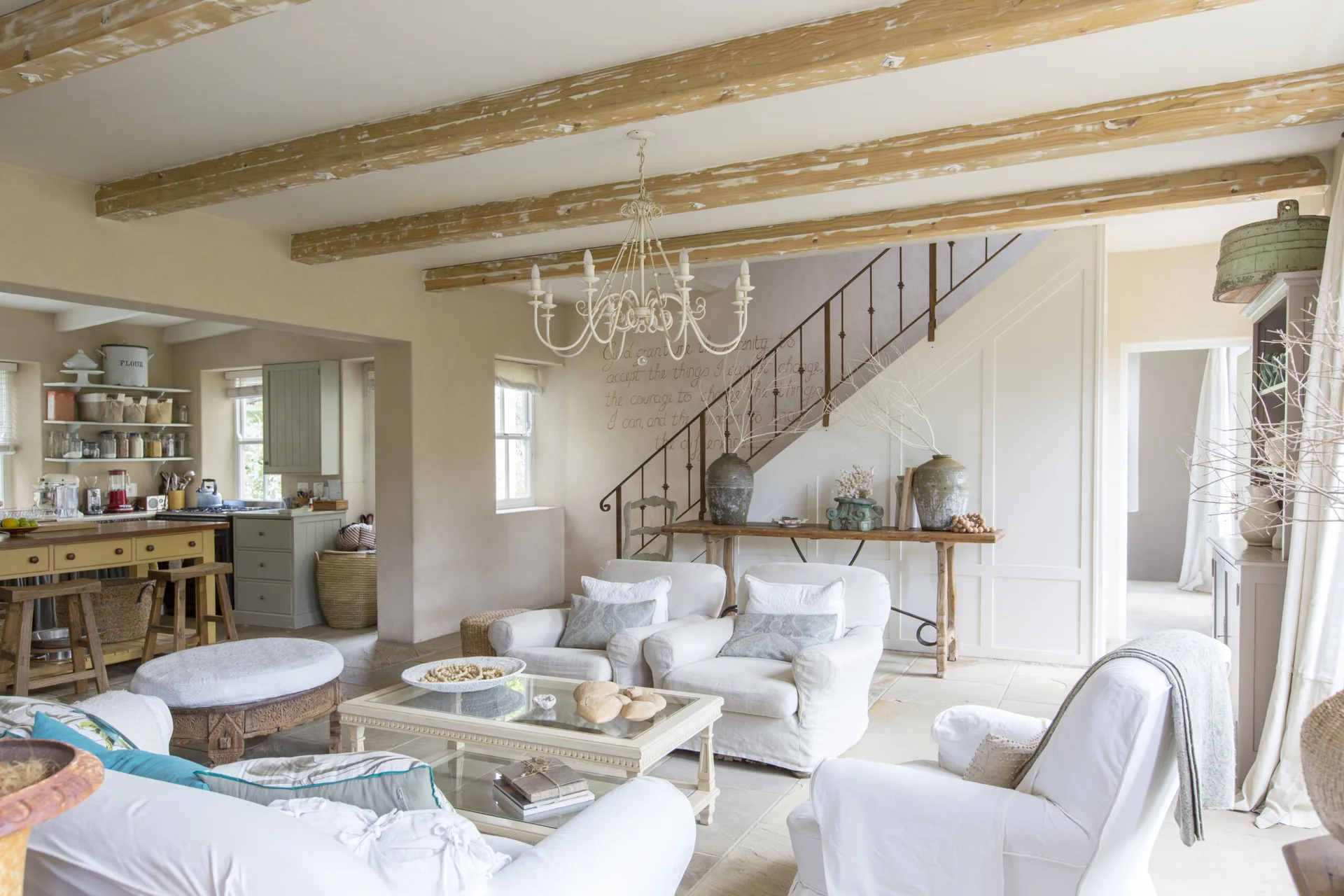 A bright farmhouse living room with white slipcovered armchairs, distressed wooden ceiling beams, a glass coffee table, and an adjacent kitchen.