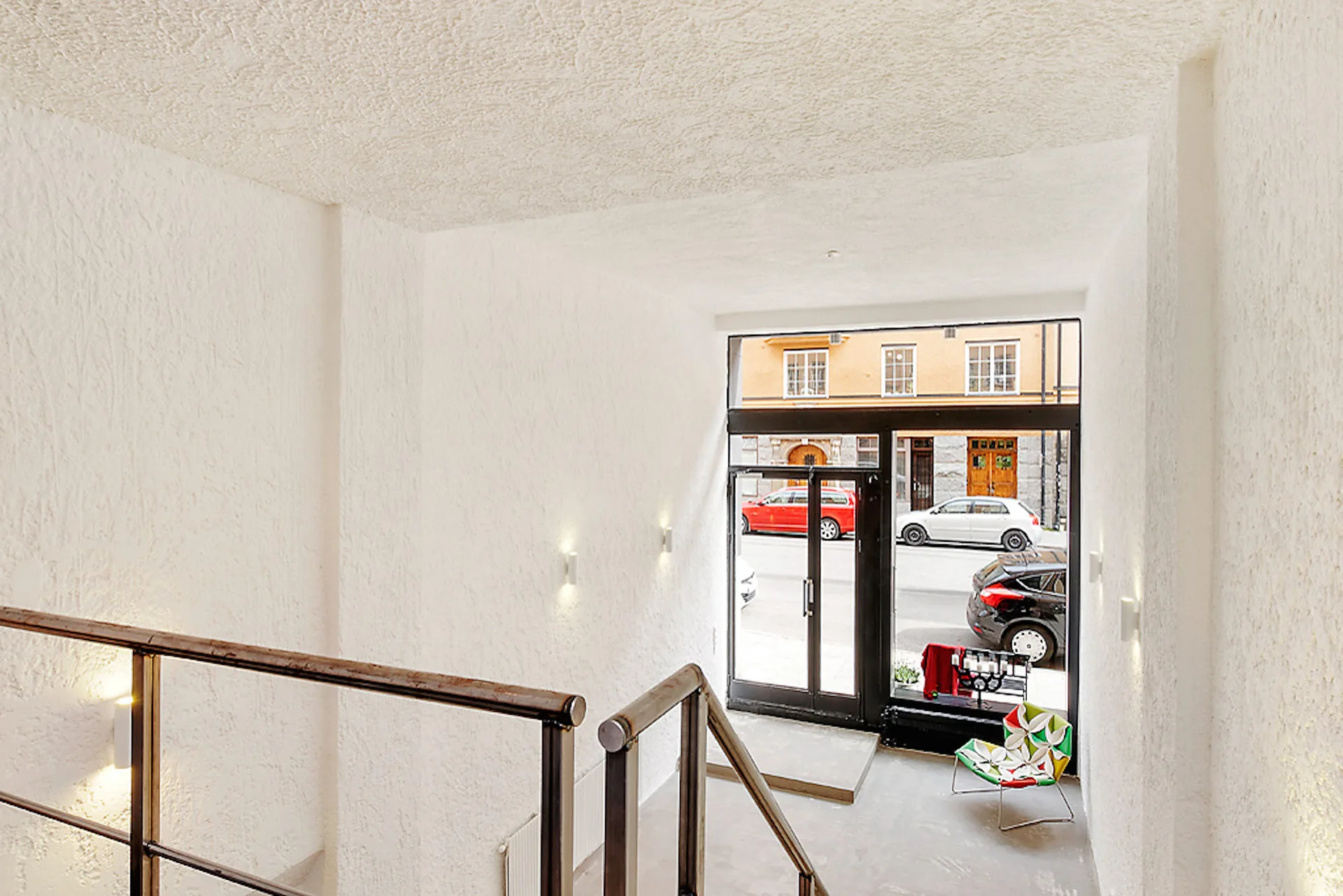 A minimalist white foyer with textured plaster walls, a dark metal staircase railing, and a glass front door showing a city street.