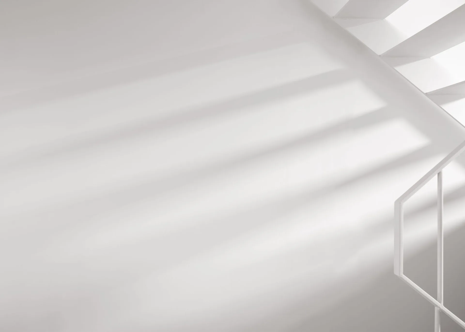 A minimalist white staircase corner features a simple metal railing and parallel shadows cast by natural light across a matte white wall.