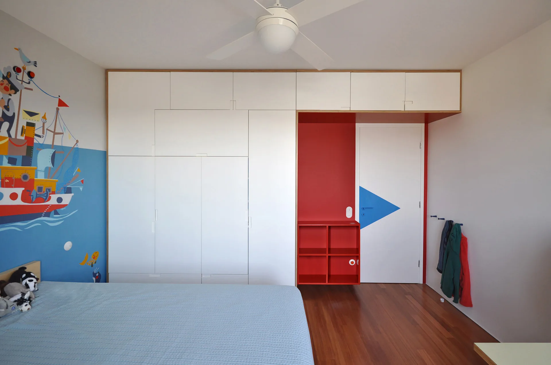 A child's bedroom featuring floor-to-ceiling white custom cabinets, an integrated red open shelving unit, and a blue nautical wall mural.