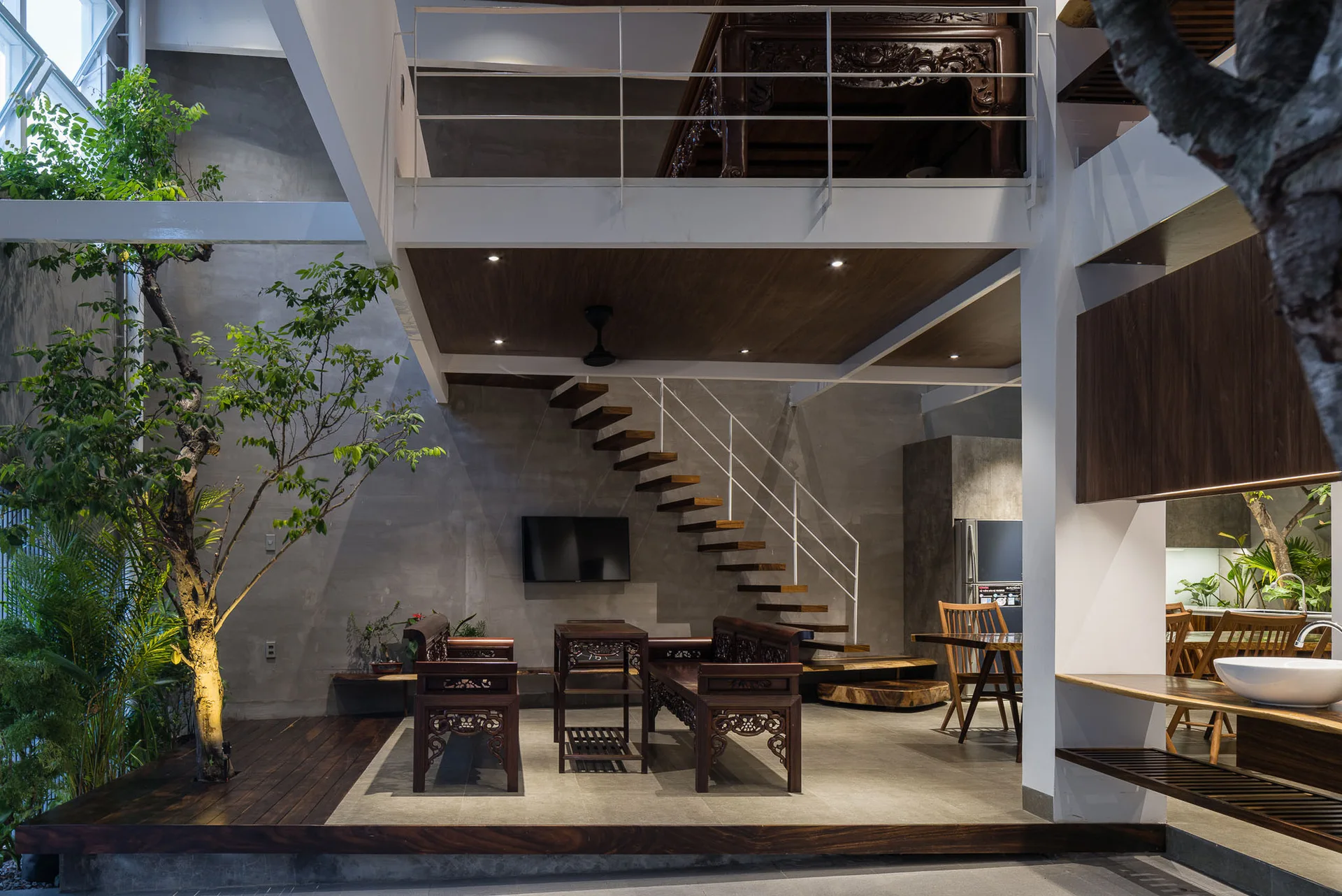An open-plan interior with raw concrete walls, a floating wooden staircase, traditional carved wood seating, and a live indoor tree.