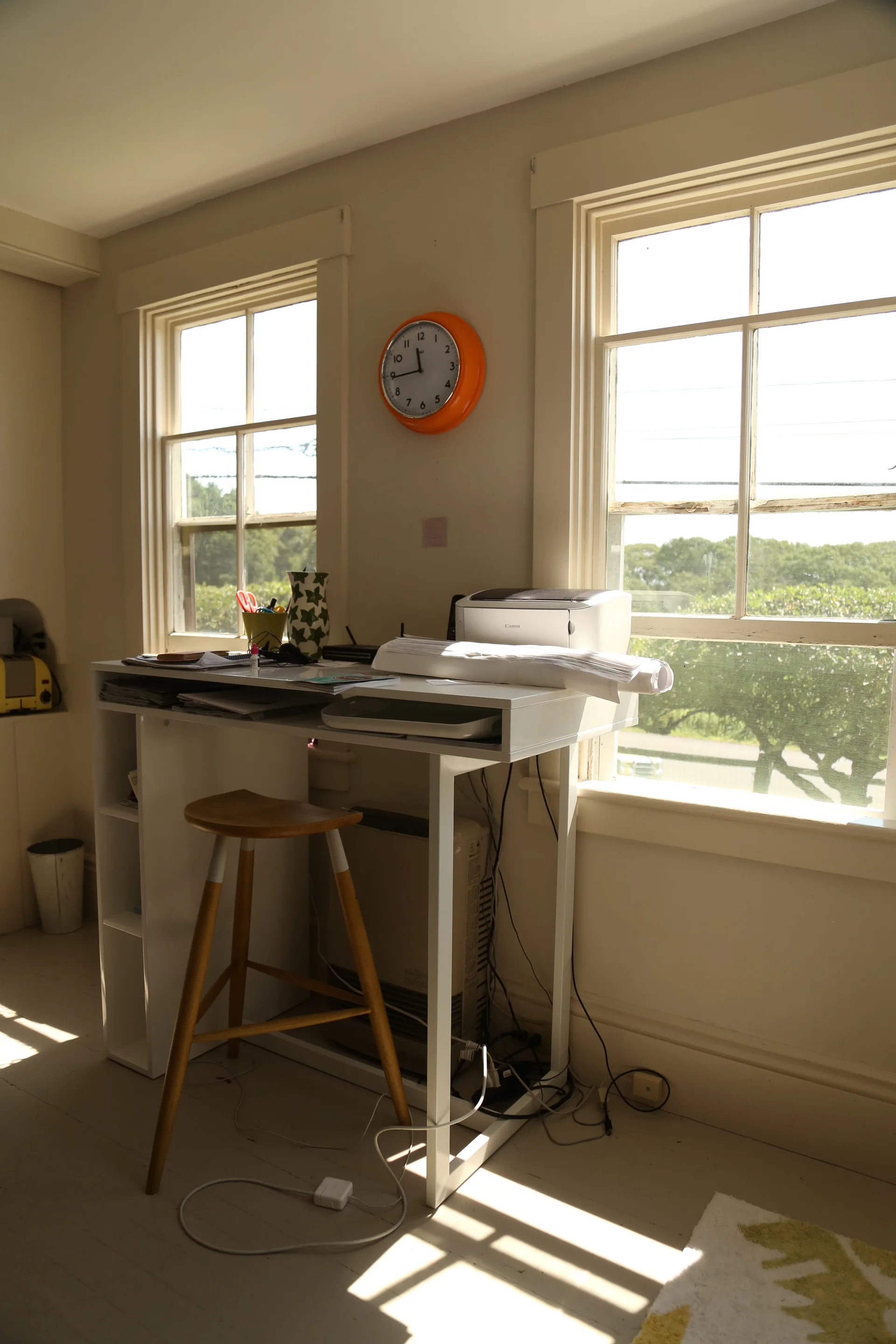 A white standing desk with a printer sits between two windows in a sunlit room, paired with a wooden stool and an orange wall clock.