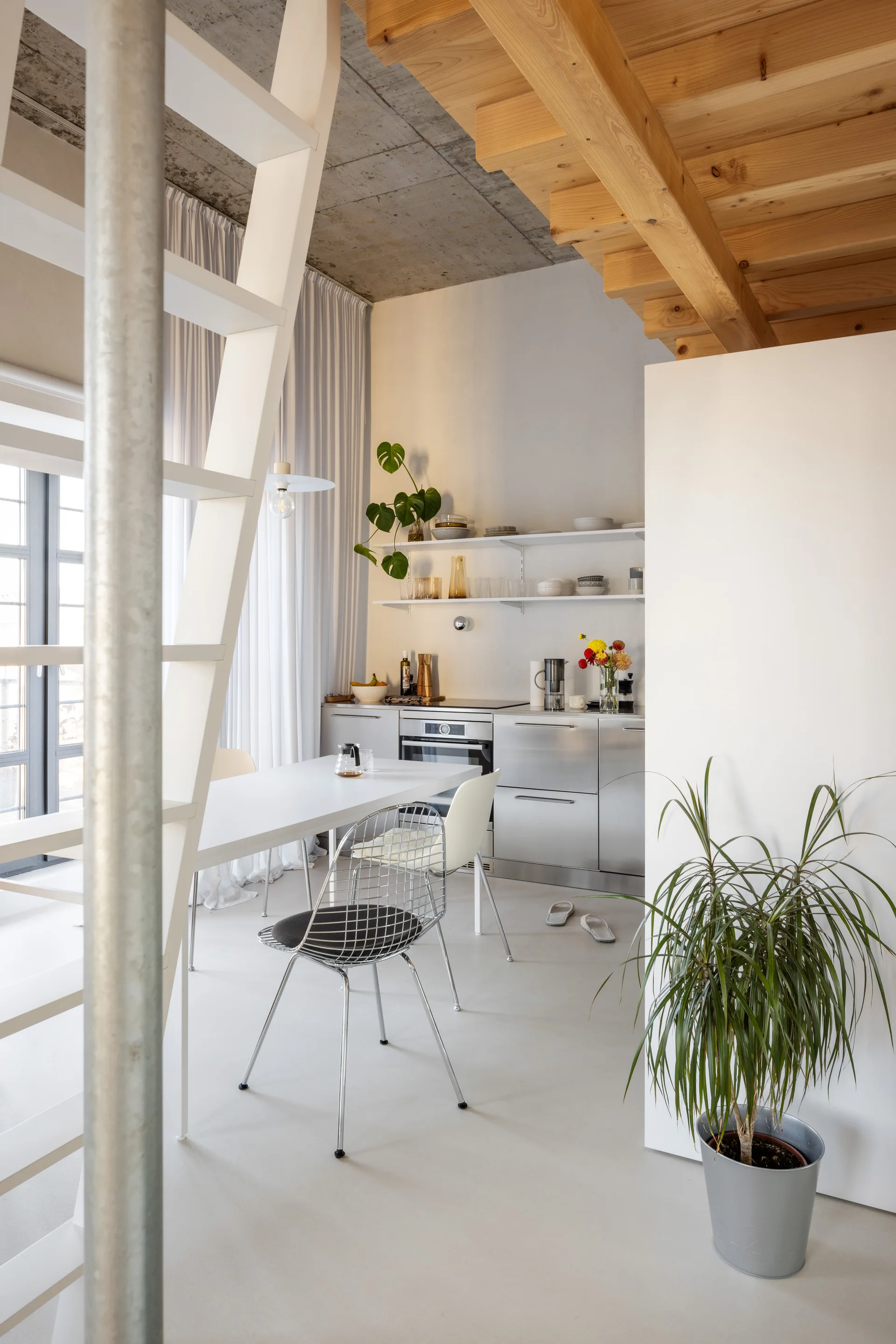 A minimalist loft kitchen featuring industrial stainless steel cabinets, a white dining table with wire chairs, and a white metal ladder.