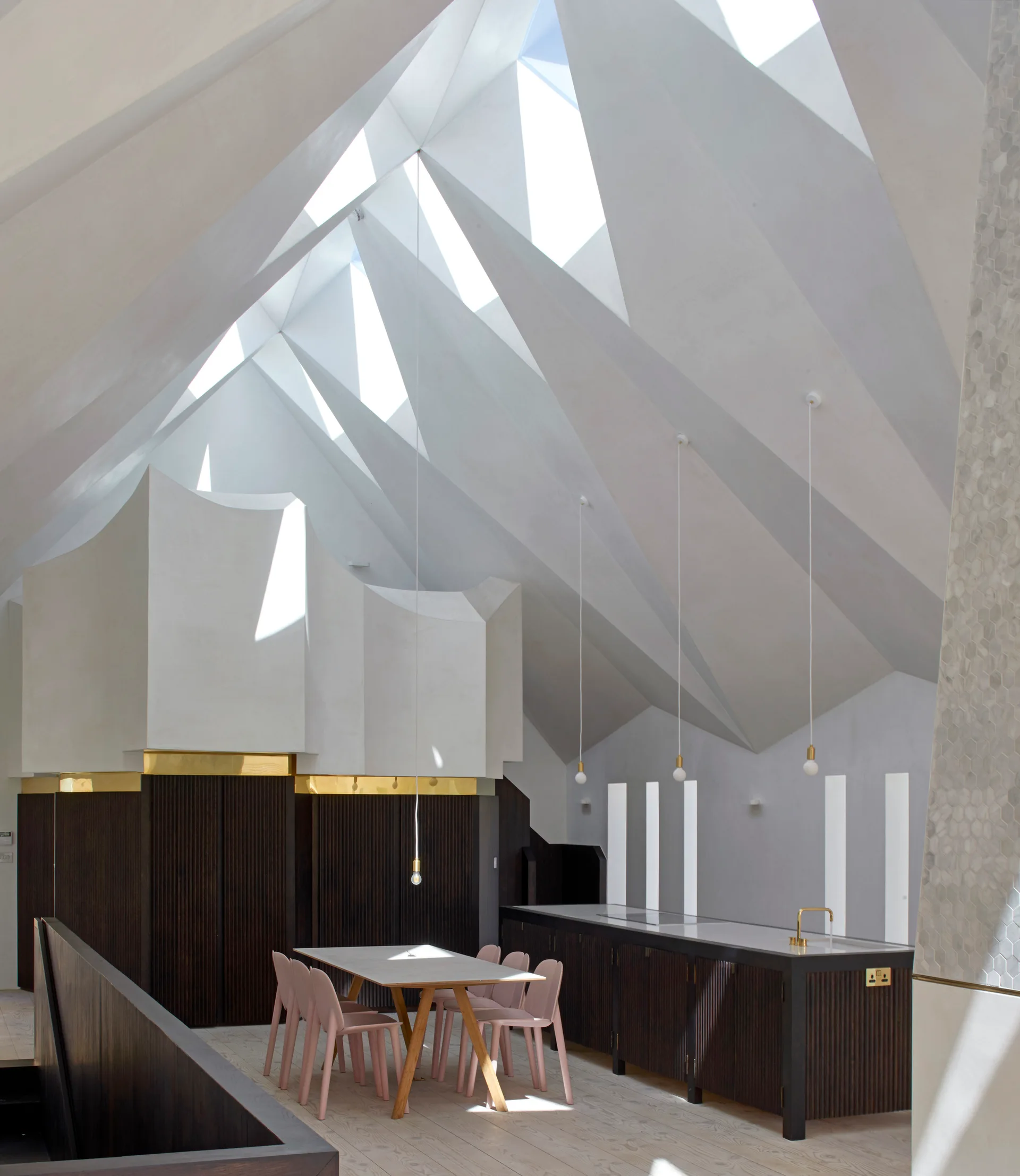 A modern kitchen and dining space with light pink chairs, a dining table, and dark fluted cabinets under a geometric white vaulted ceiling with skylights.