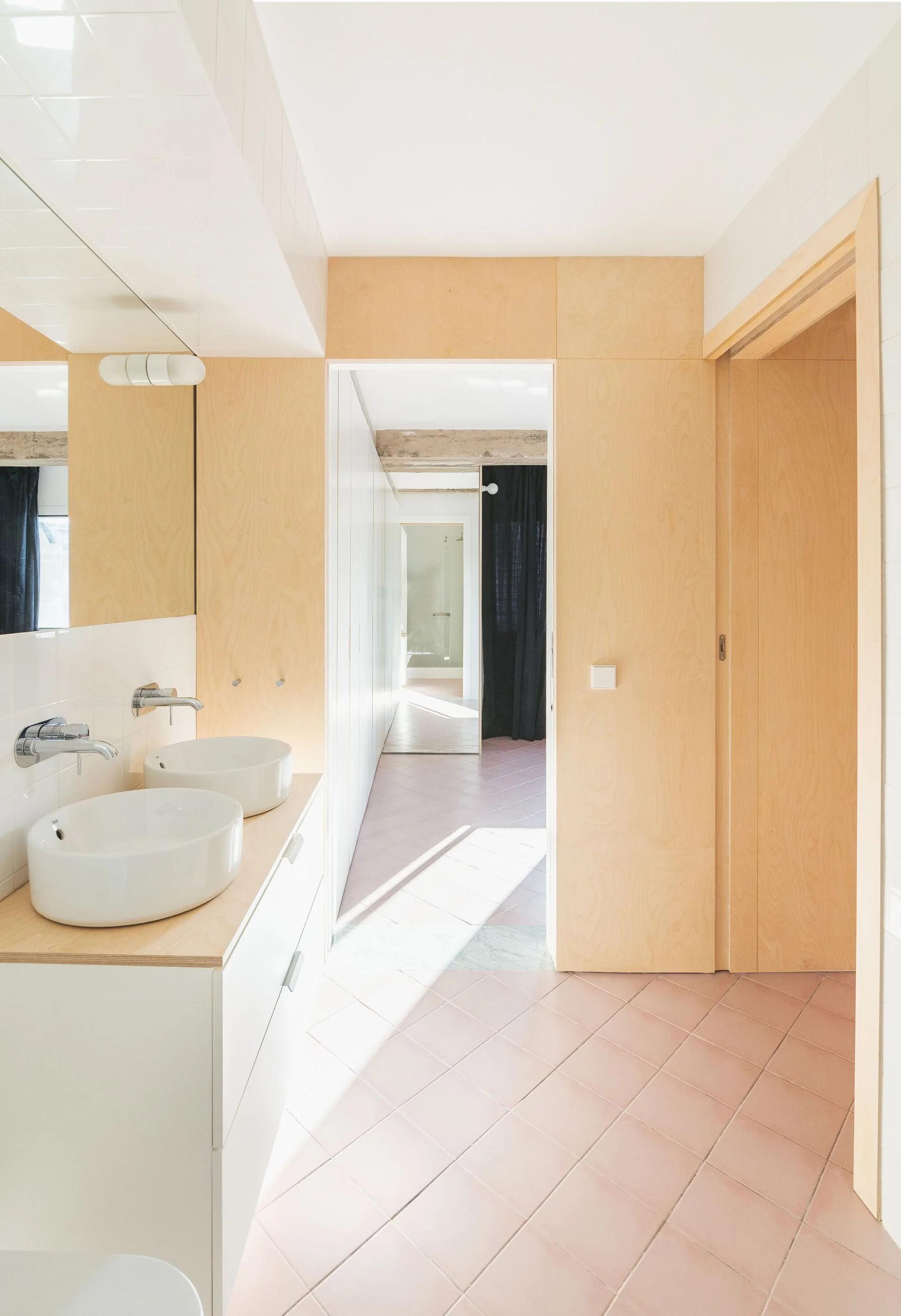 A minimalist bathroom with a double vessel sink vanity, light birch plywood wall panels, and pink square floor tiles.