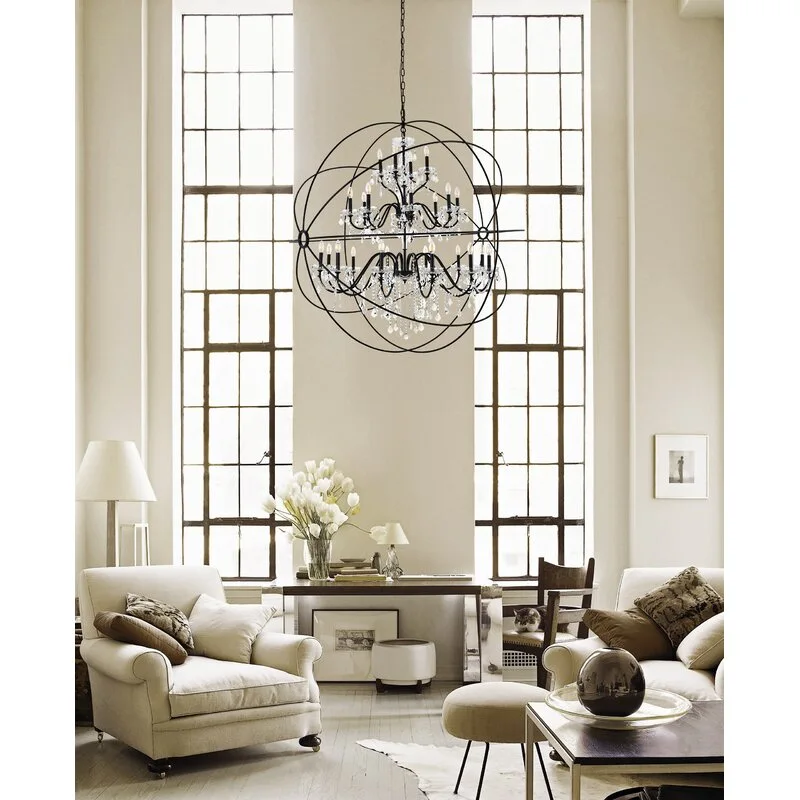 A bright living room with cream armchairs, two tall paned windows, and a large black metal orb chandelier hanging from the high ceiling.