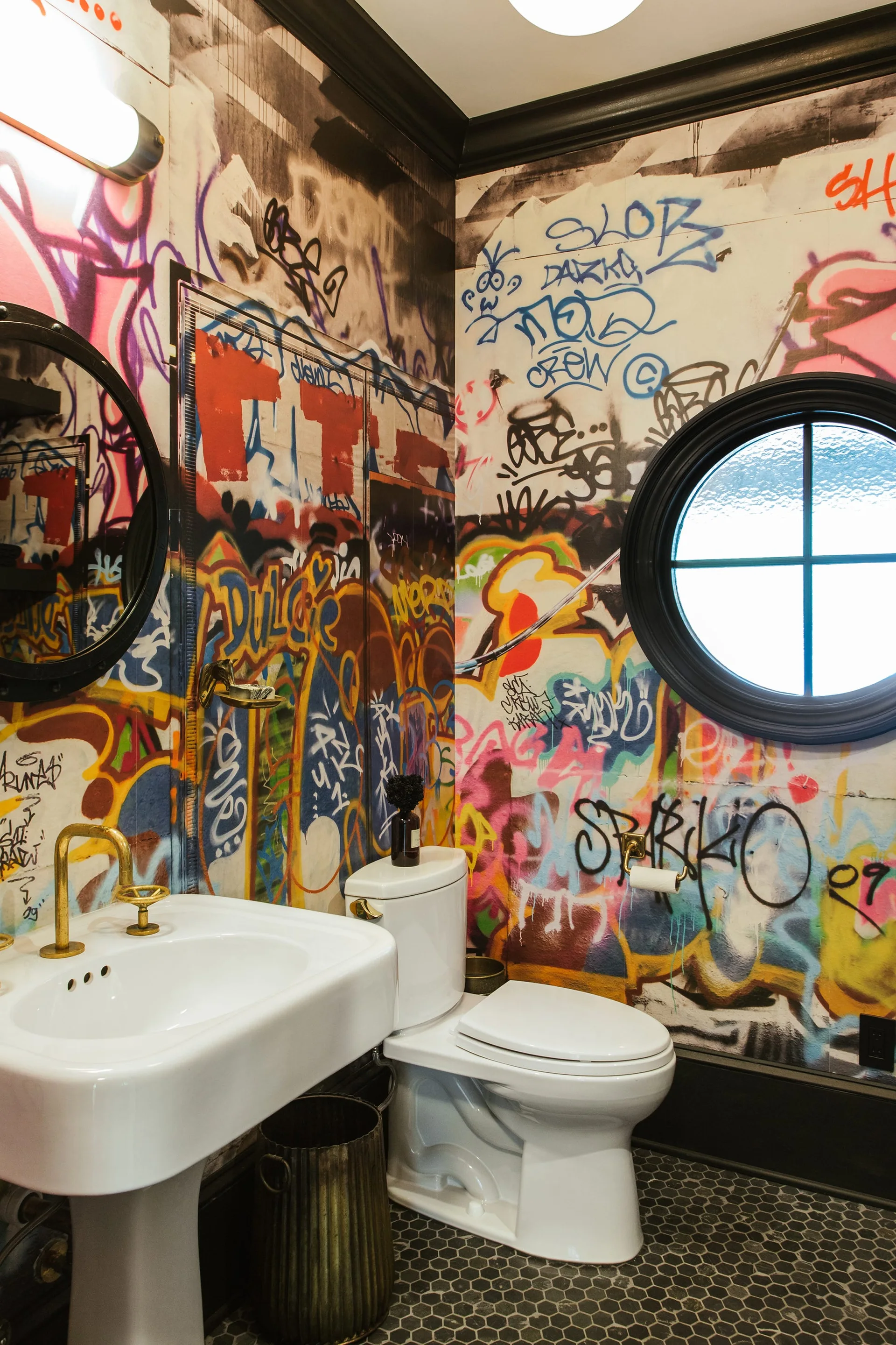A powder room features colorful graffiti wallpaper, a white pedestal sink with a gold faucet, a toilet, and a circular black-framed window.