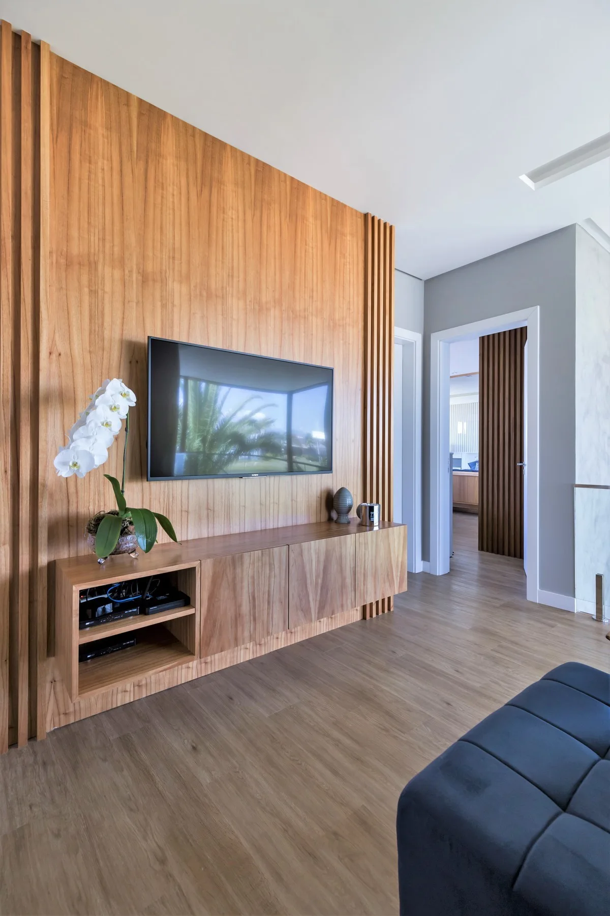 A modern living room features a vertical wood slat feature wall with a wall-mounted TV, a floating wooden media console, and a white orchid.