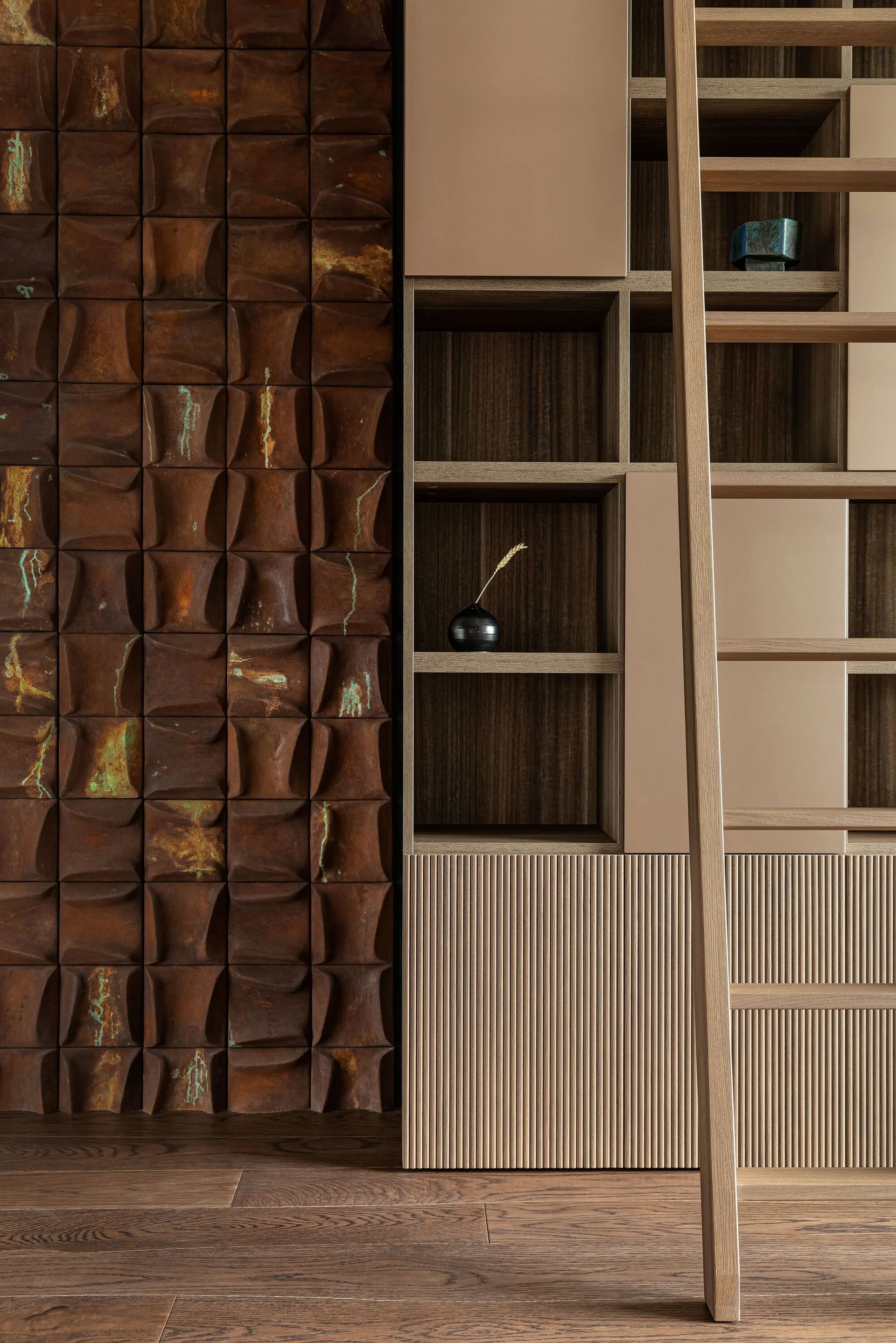 A modern wooden shelving unit with a library ladder stands next to a wall clad in textured, rust-colored square metal panels with green patina.
