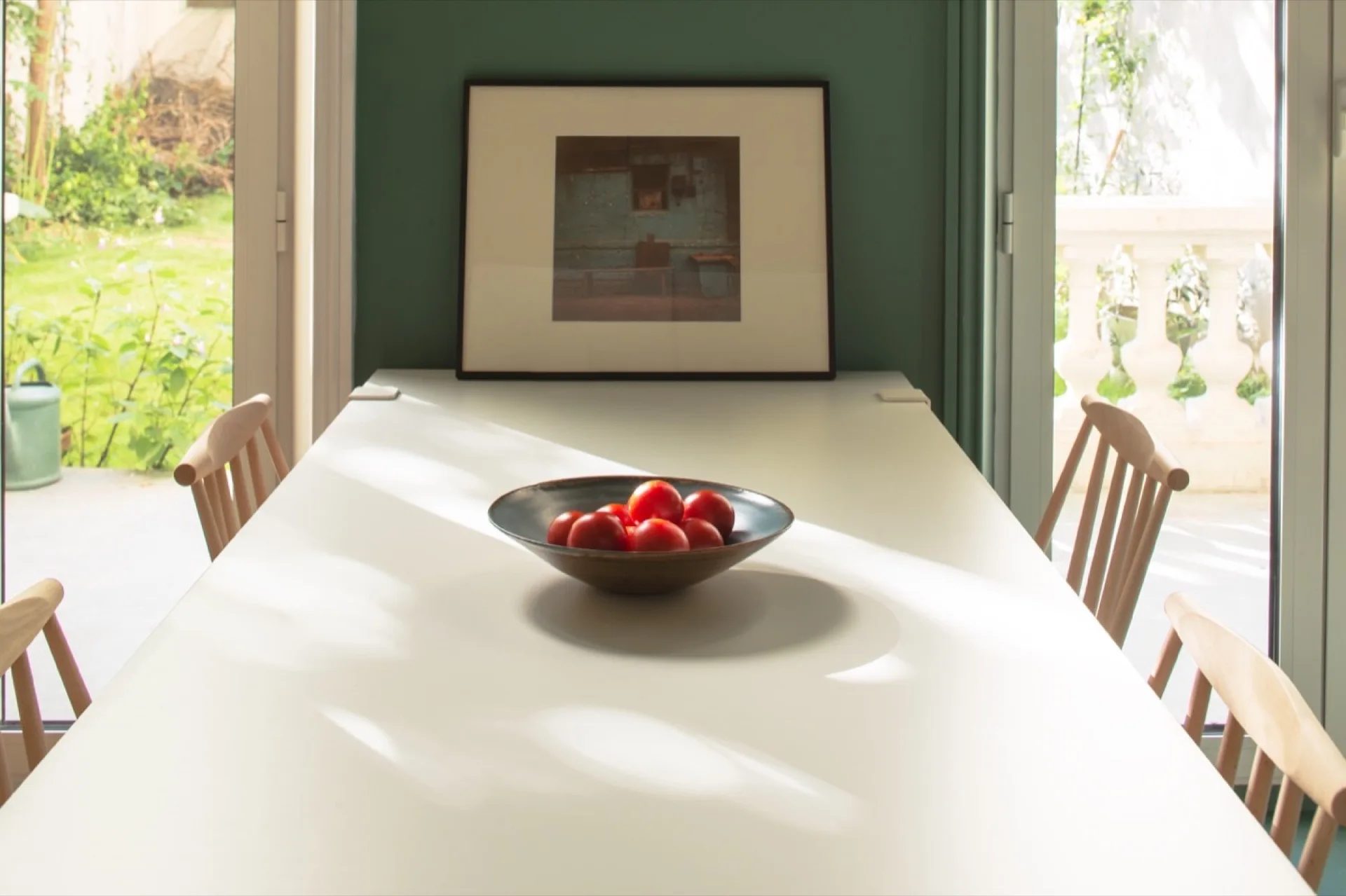 A white rectangular dining table with light wood spindle-back chairs and a ceramic bowl of red tomatoes, set against a dark green wall with a framed print.