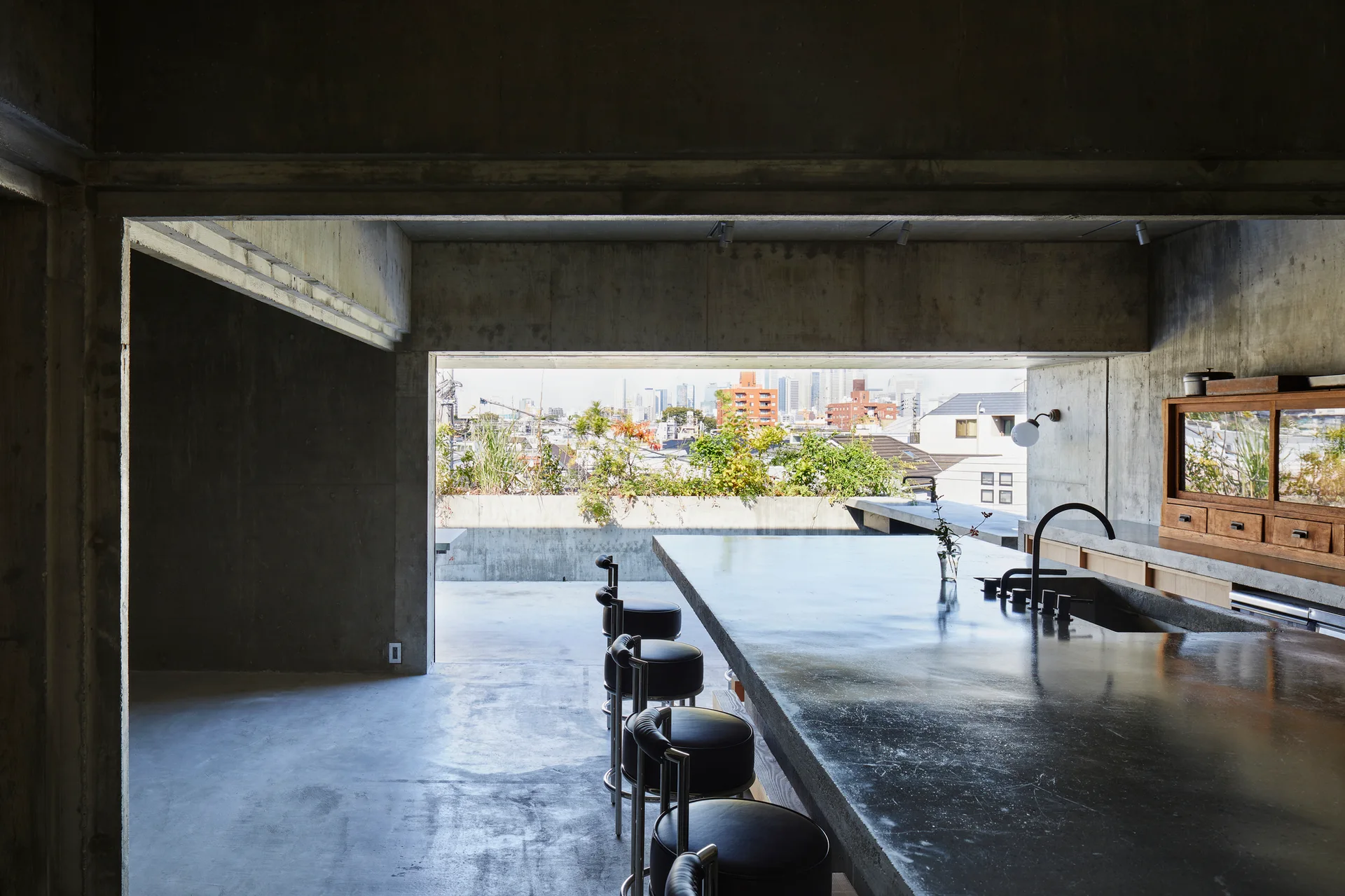 An industrial kitchen features exposed concrete walls, a large island with black bar stools, and an open entryway leading to an outdoor balcony.