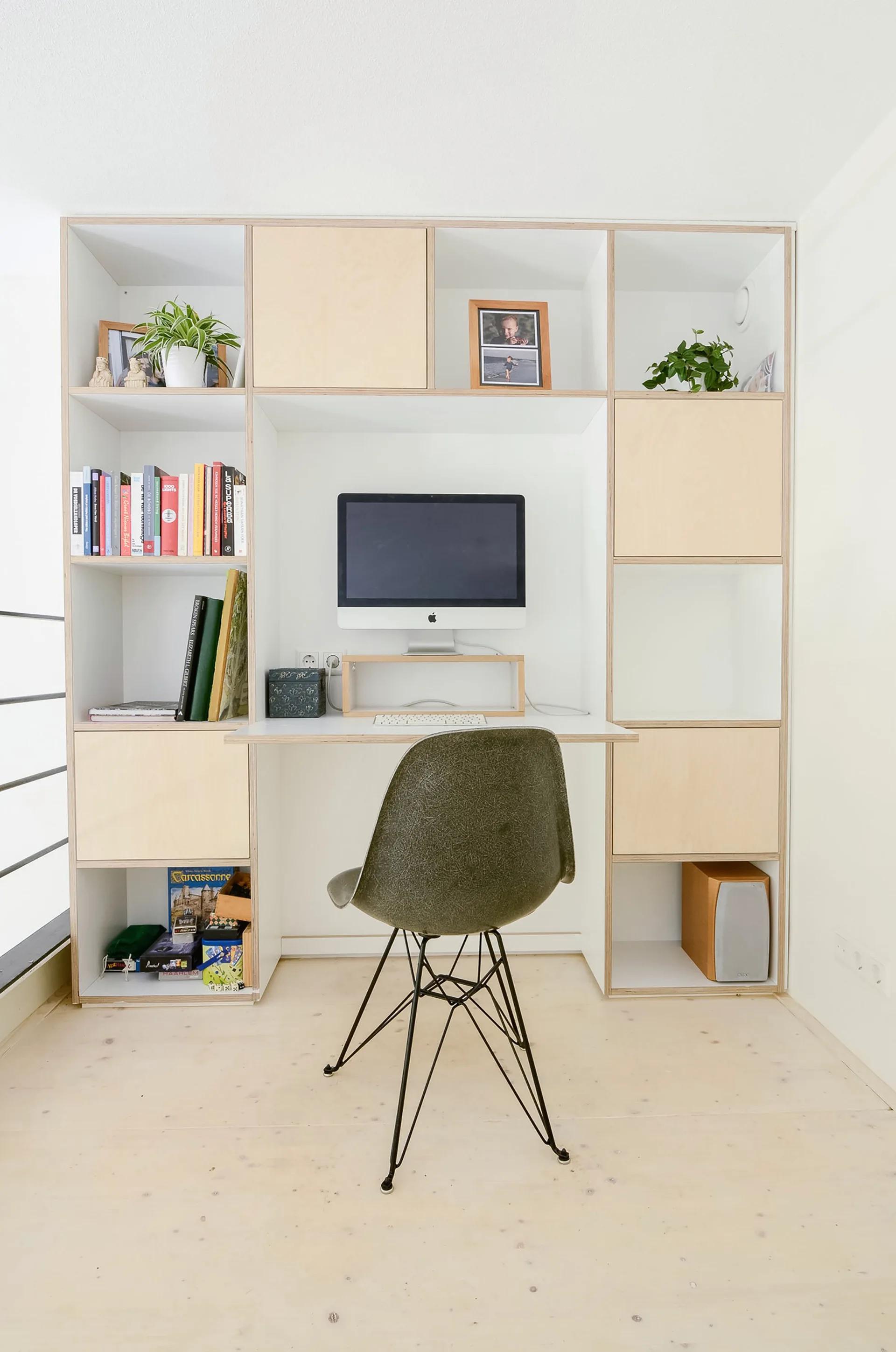 A custom plywood shelving unit with an integrated white desk features an olive green molded chair, a desktop computer, and organized books.