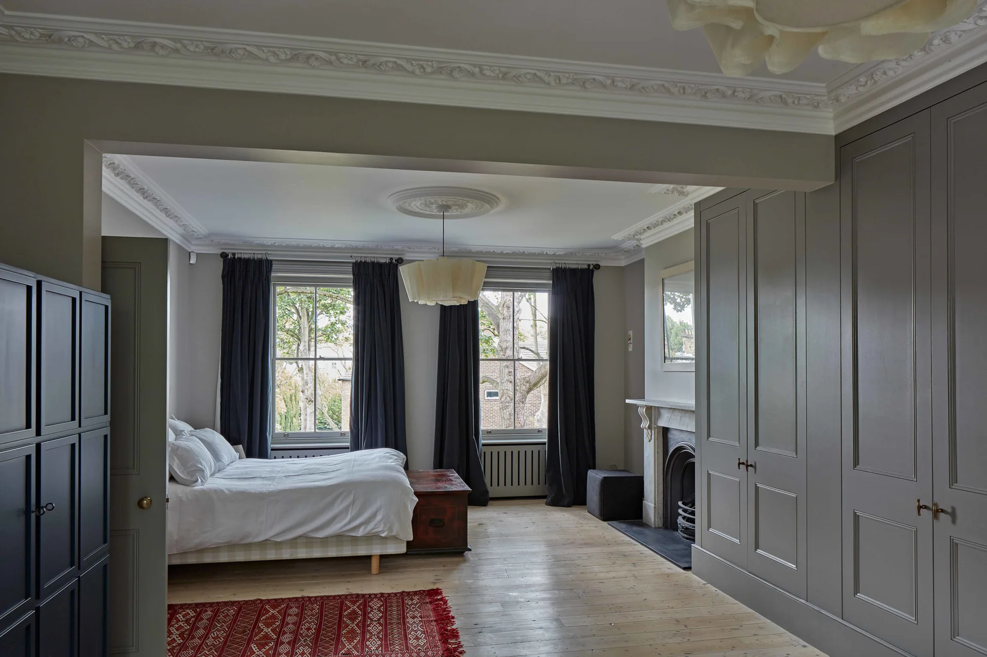 A classic bedroom featuring a white bed, tall grey built-in wardrobes, sash windows with dark curtains, and a marble fireplace on light wood floors.