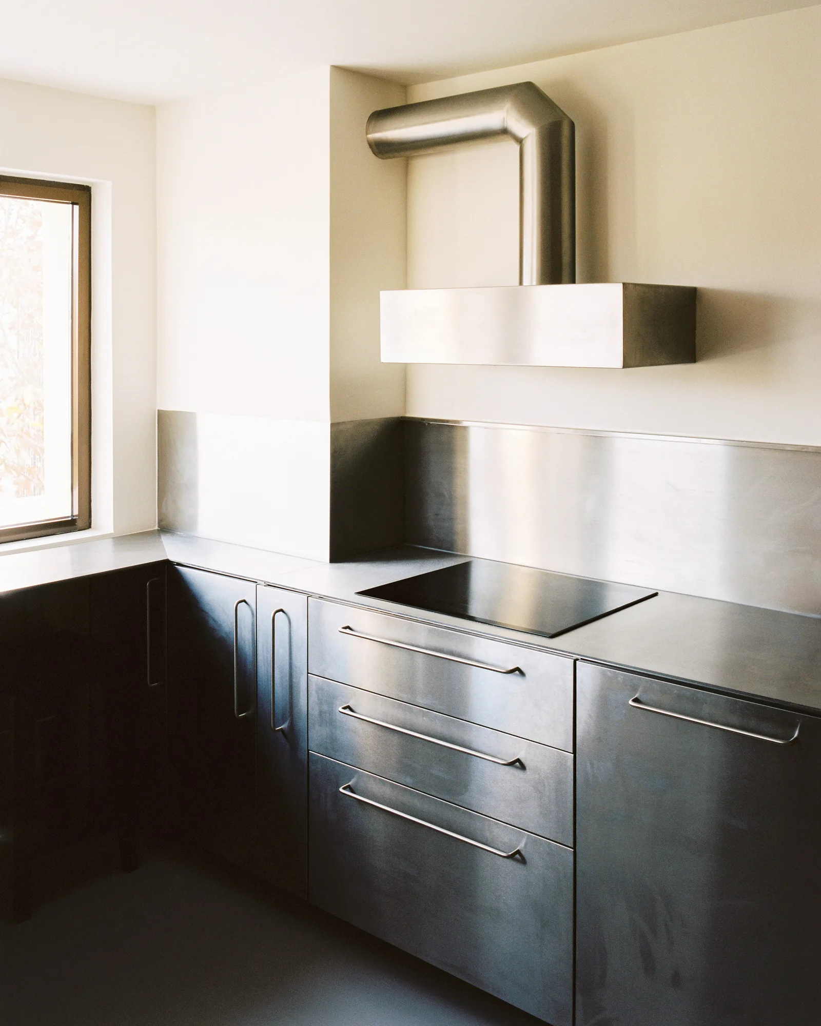 Minimalist kitchen corner featuring brushed stainless steel cabinets, a matching backsplash, an integrated cooktop, and an exposed metal range hood.