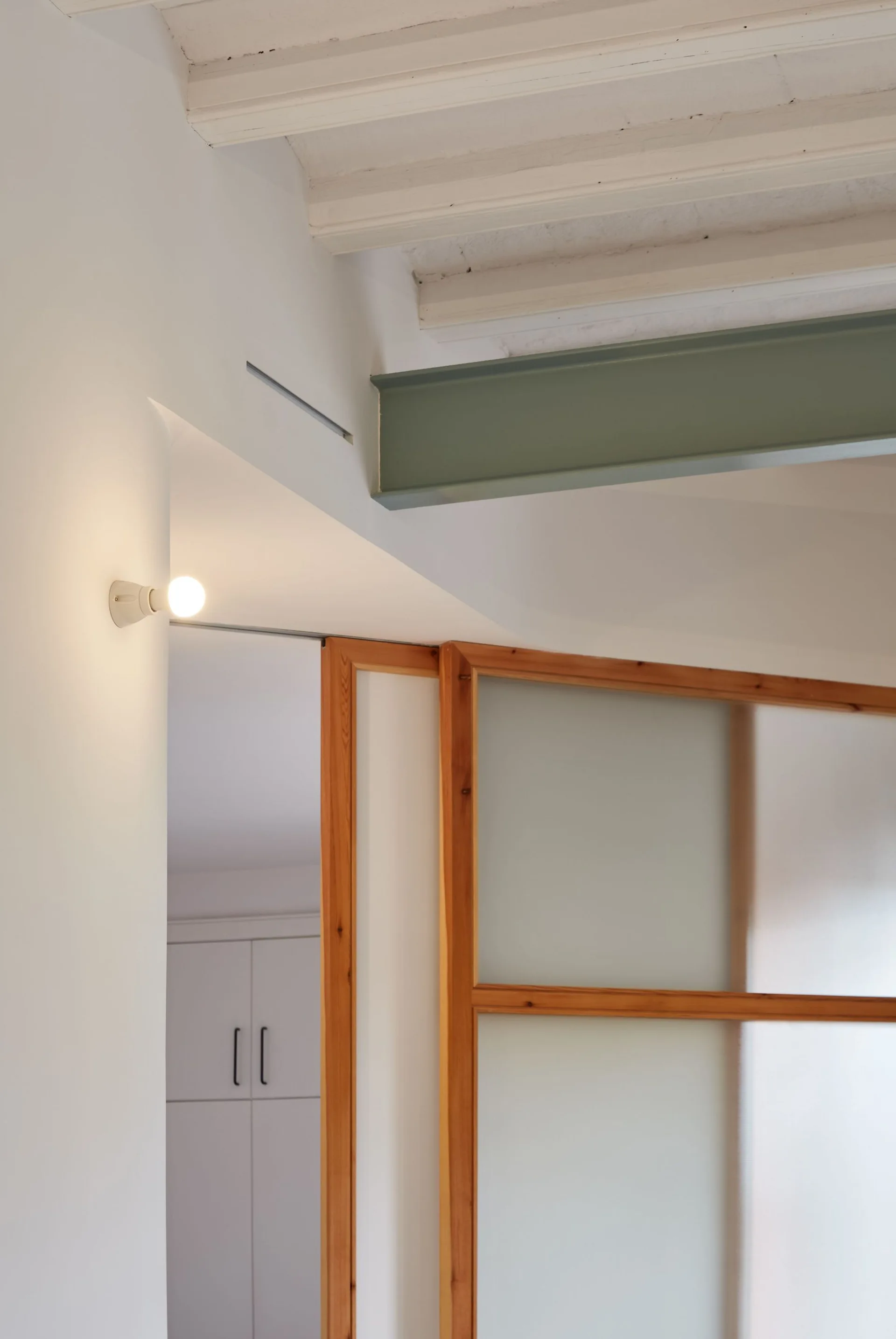 A sage green structural beam runs across a white ceiling above a wooden-framed frosted glass sliding partition and a glowing wall sconce.