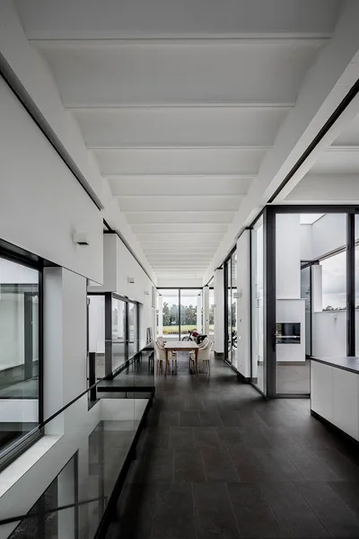 A long minimalist corridor with dark tile flooring, a white ribbed ceiling, and glass railings leading to a distant dining area with large windows.