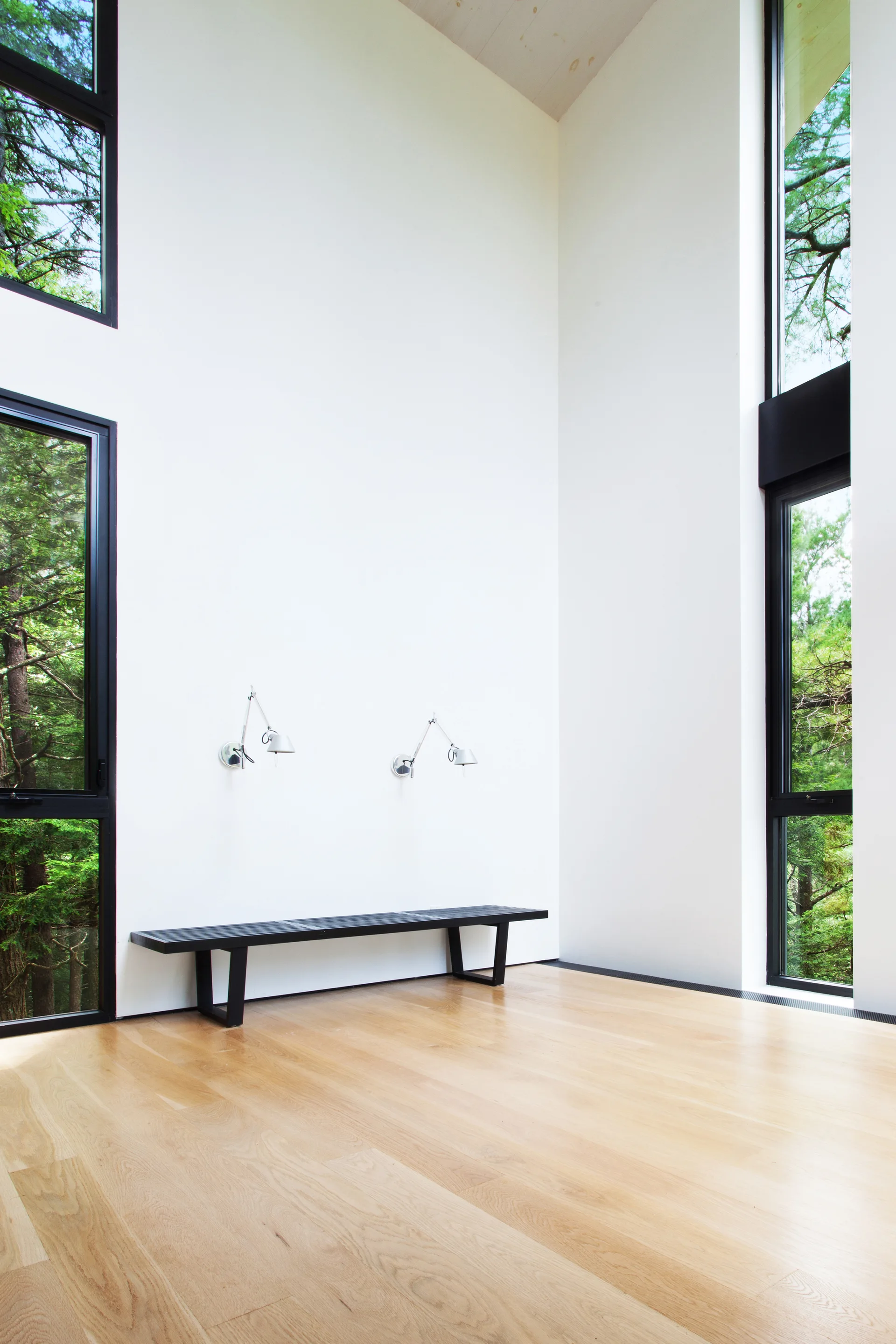 A minimalist room features light hardwood floors, high white walls, tall black-framed windows, a black slatted bench, and two silver wall sconces.