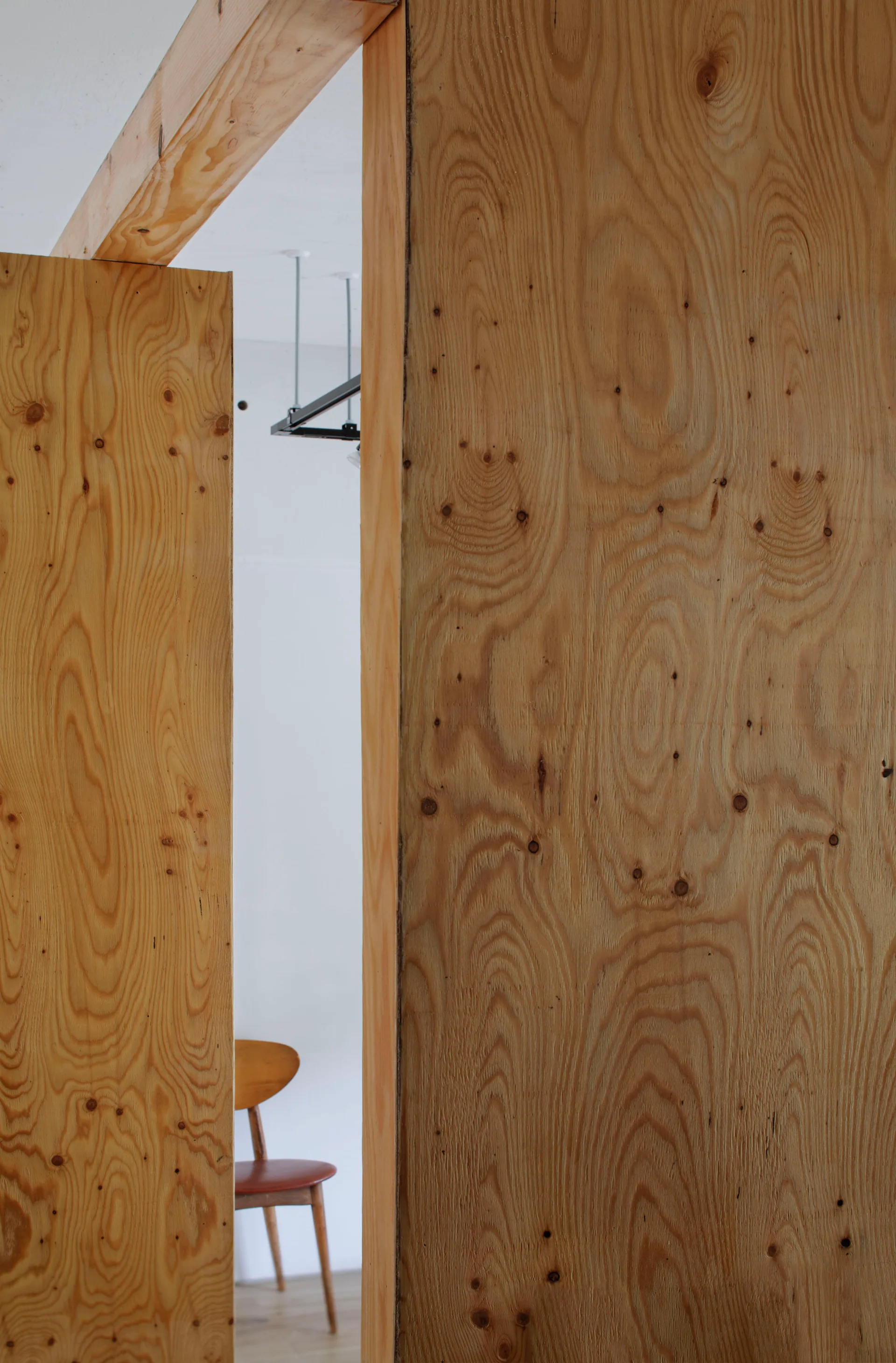 A close-up view through a plywood door frame with a prominent wood grain pattern reveals a white room with a classic wooden chair.