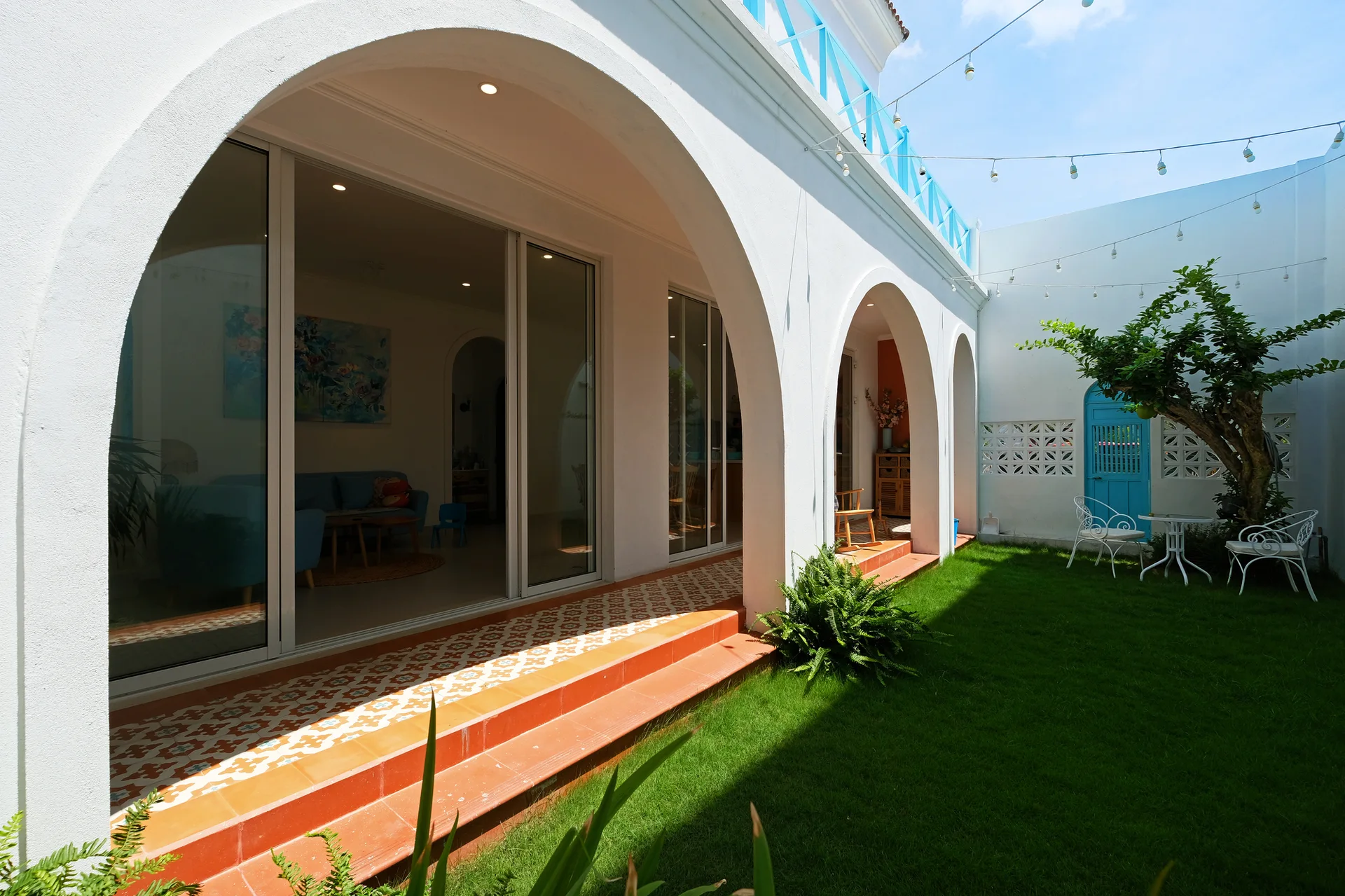 White stucco arches frame a covered porch with geometric pattern tiles next to a green grass lawn with metal garden furniture.