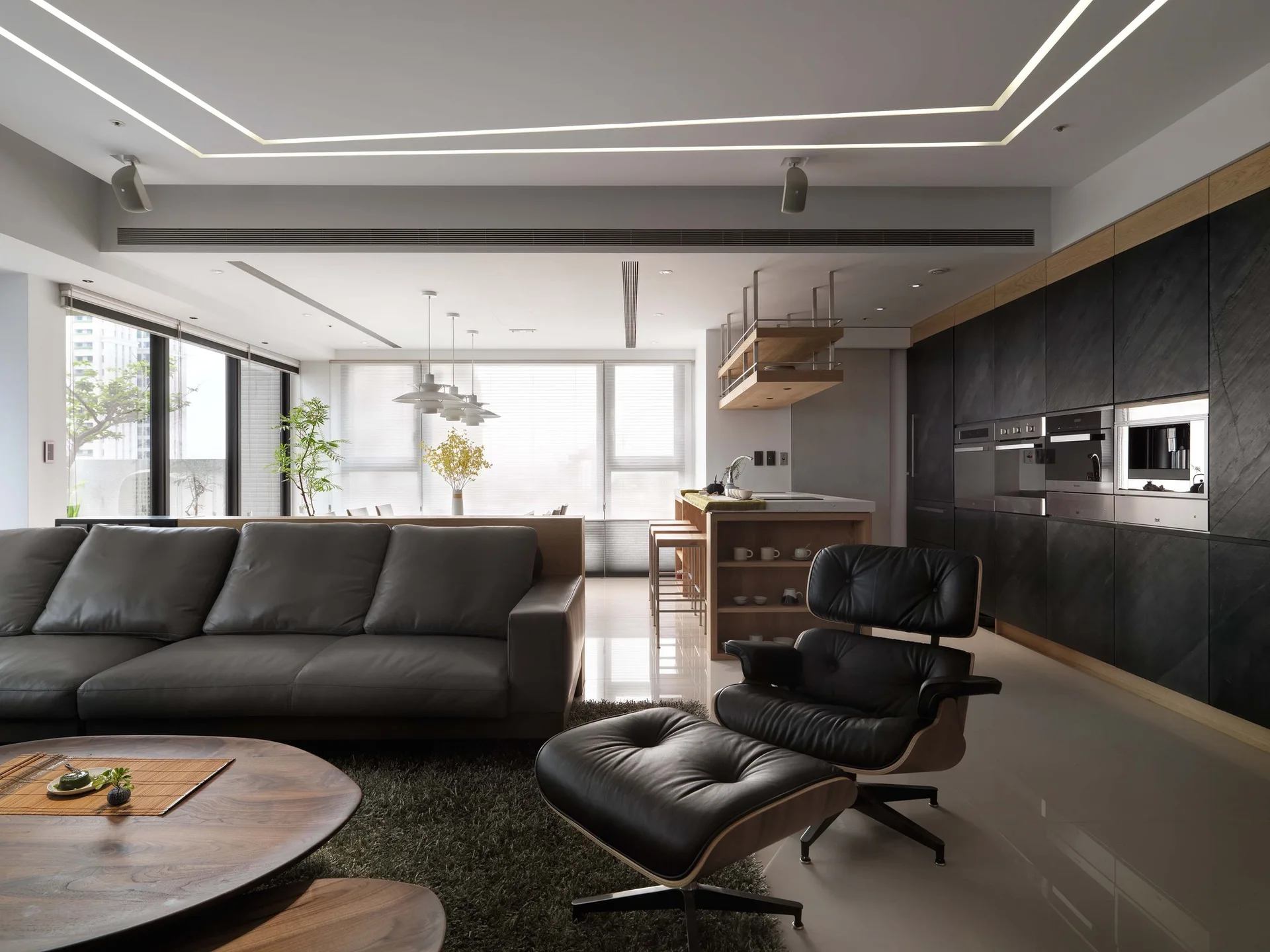 A modern open-plan living space features a dark grey leather sofa, a classic black leather lounge chair, and a matte black kitchen cabinet wall.