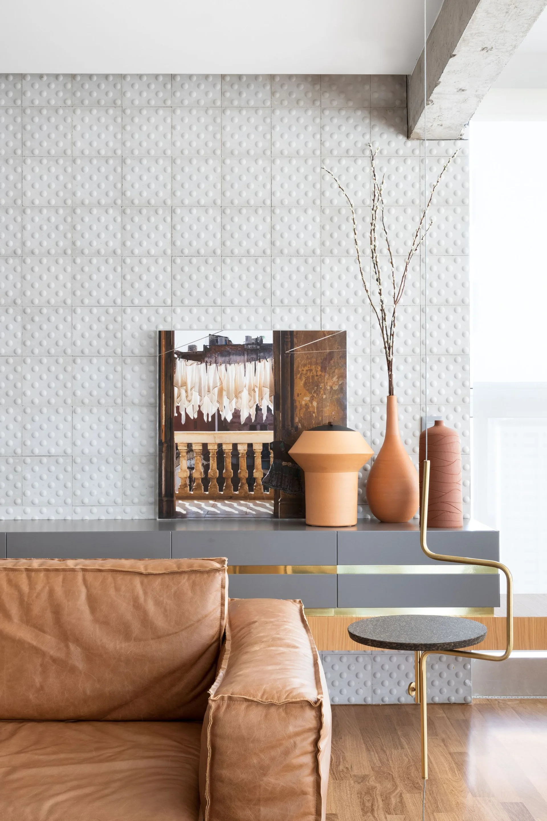 A brown leather sofa sits in front of a grey credenza styled with terracotta vases and artwork against a white textured dot wall.