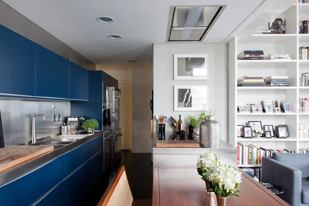 A modern kitchen featuring matte blue cabinets, a stainless steel backsplash, and a tall white built-in bookshelf filled with books and frames.