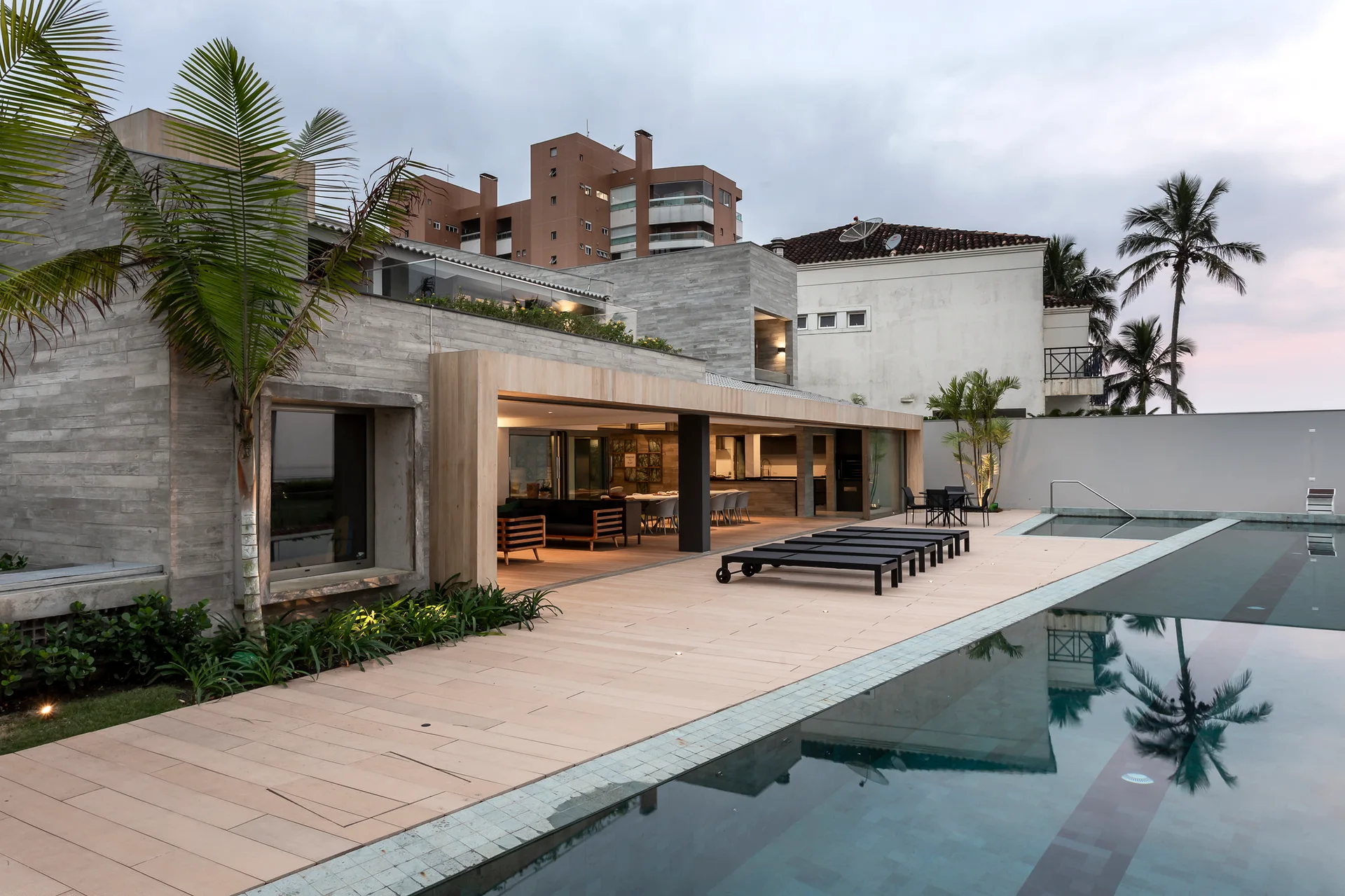 A modern grey concrete house features a wide wood-plank deck, black lounge chairs beside a swimming pool, and an open-plan indoor living space.