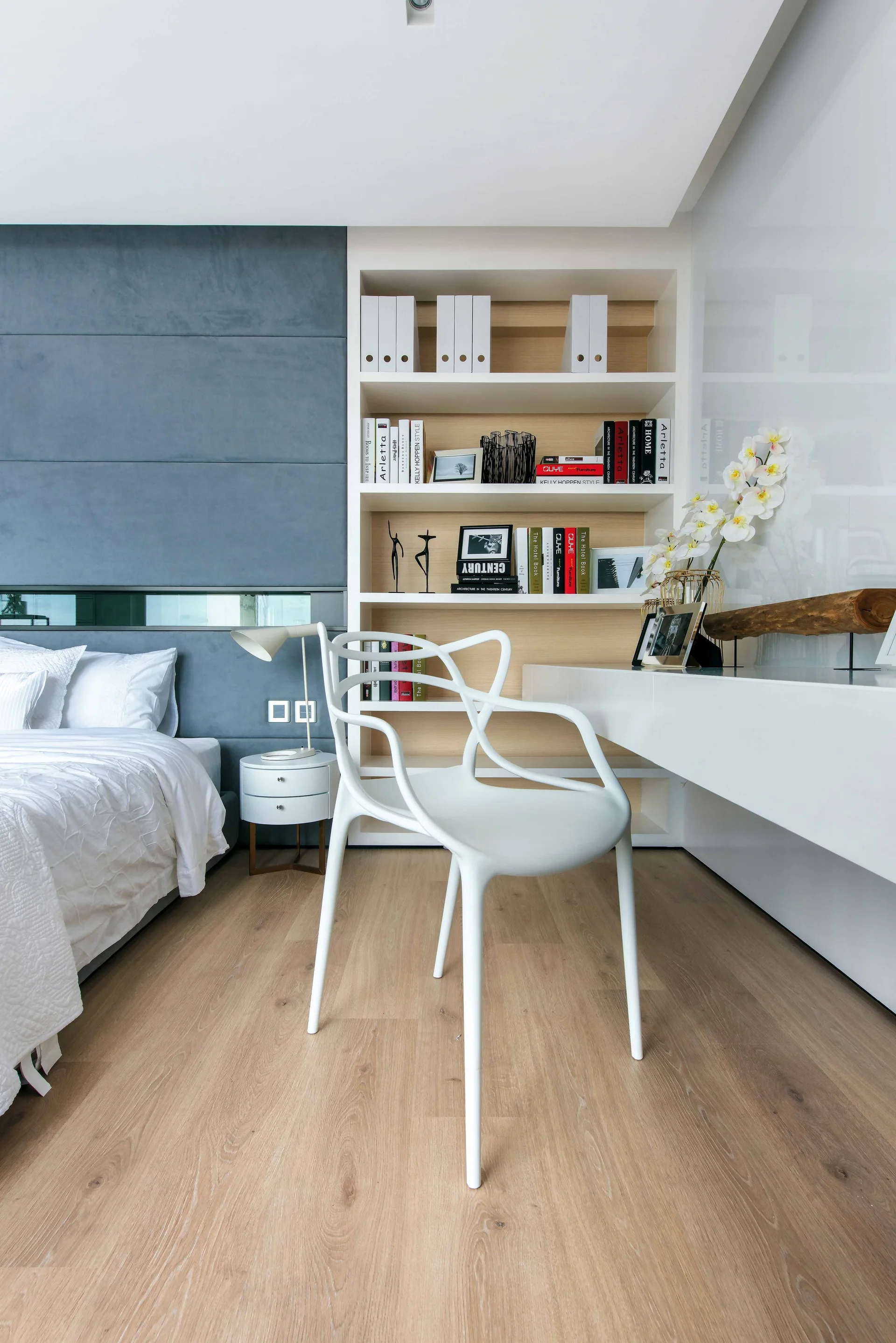 A modern bedroom features a blue upholstered wall panel behind the bed, a white molded chair, and an integrated desk with open wooden shelving.