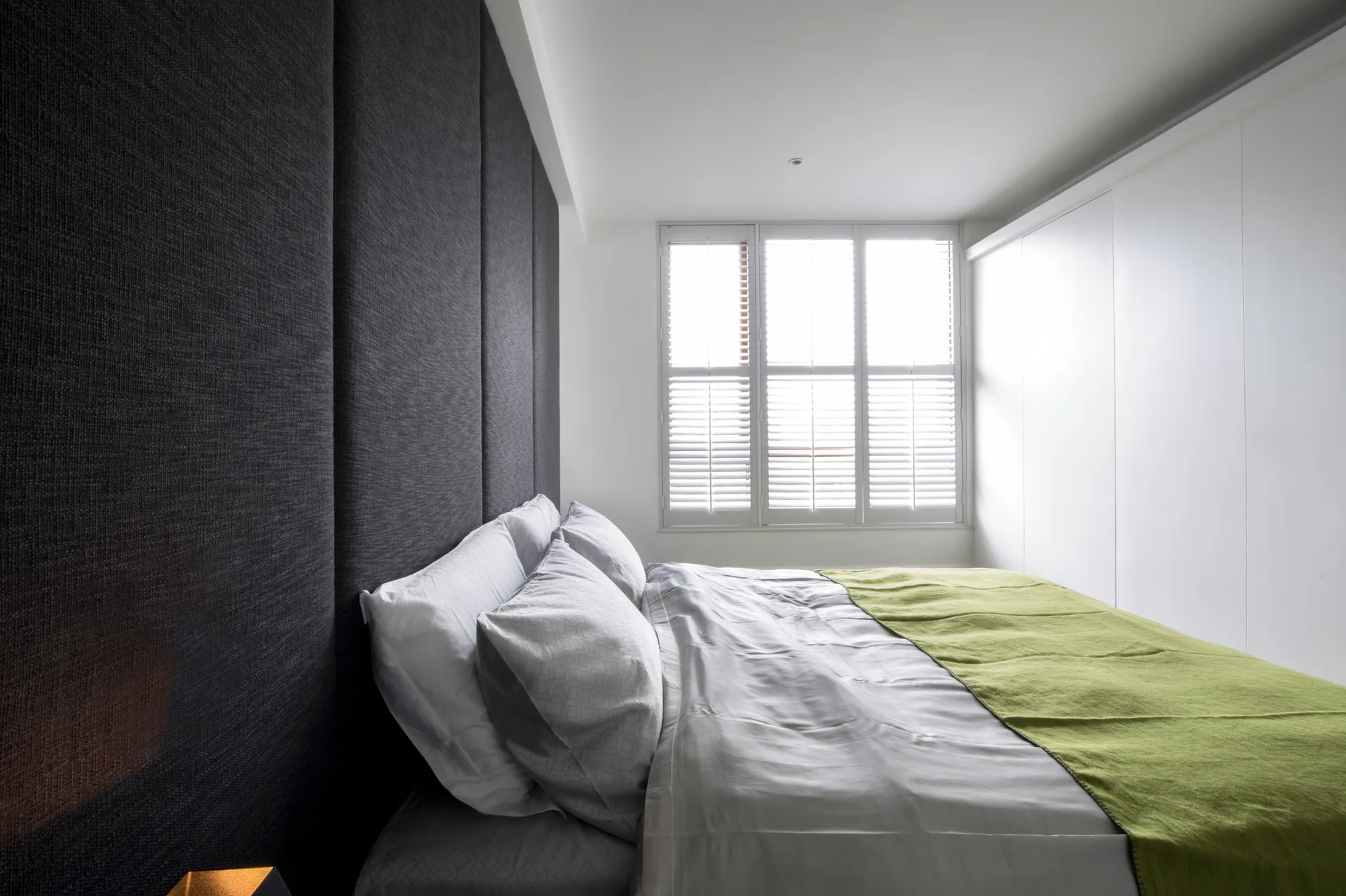 A modern bedroom features a full-height dark grey fabric paneled wall behind a bed, white plantation shutters, and white built-in closets.