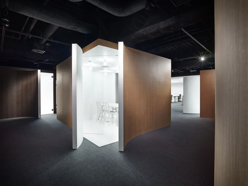A modern office interior featuring a freestanding meeting pod with curved wood-toned walls and a bright white glass interior.