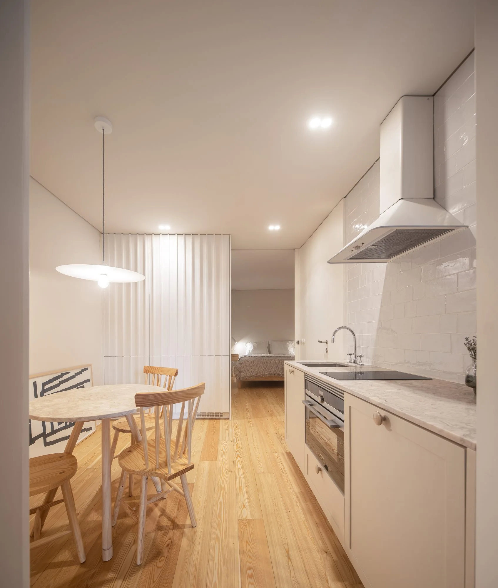 A compact modern kitchen with off-white cabinets faces a small round dining table, with a slatted partition wall leading to a bedroom.