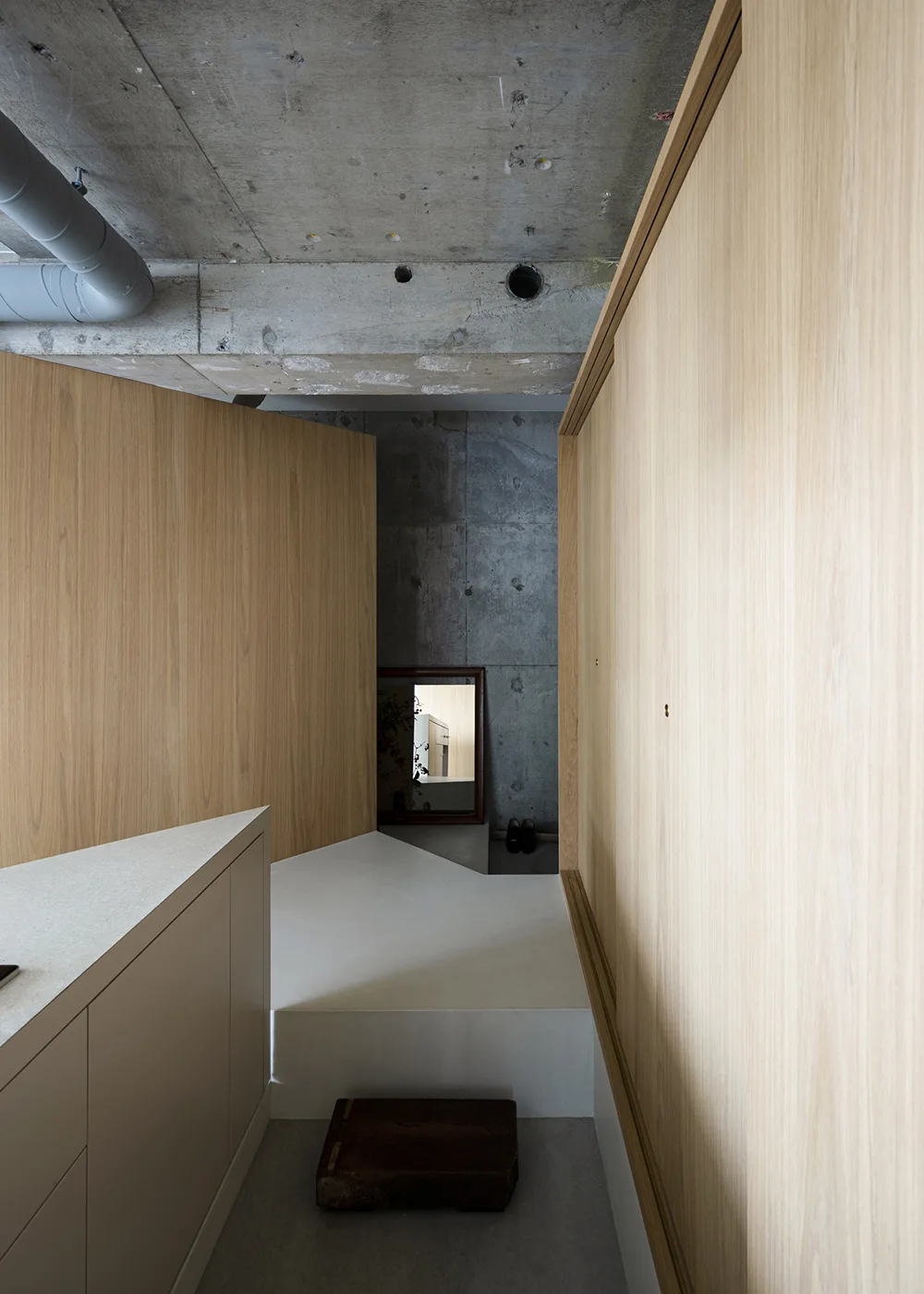A minimalist entryway features light wood-paneled walls, sliding cabinetry doors, a platform floor, and an exposed raw concrete ceiling.