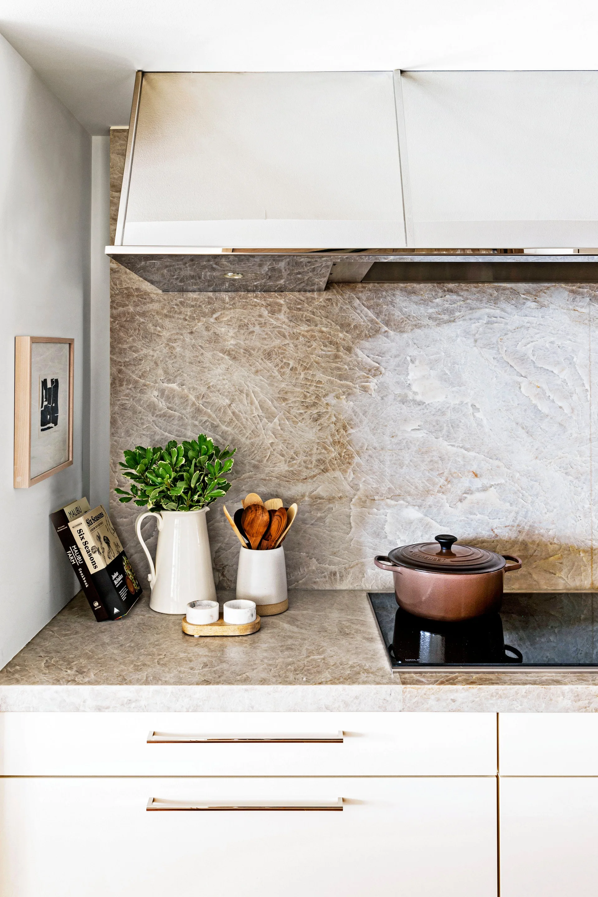 A kitchen counter features a beige veined stone backsplash and countertop, white cabinetry, a black cooktop with a brown pot, and decorative kitchen items.