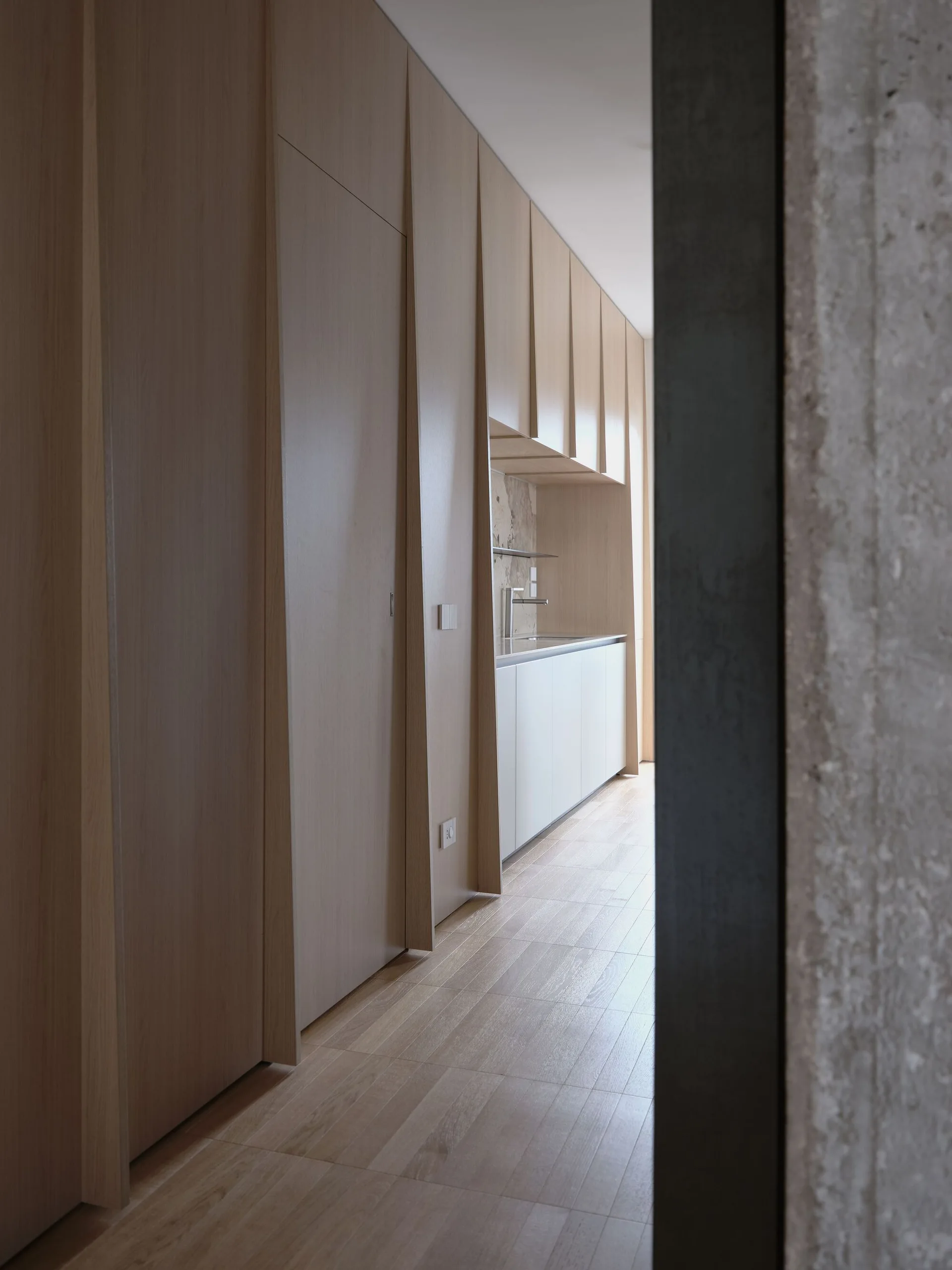 A minimalist corridor framed by a textured concrete column features integrated light wood cabinetry leading to a white kitchen counter.